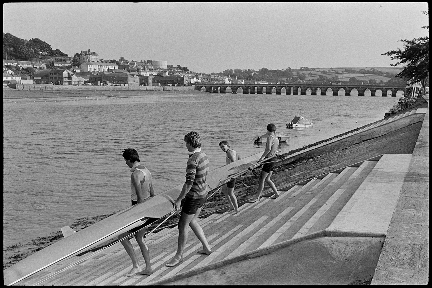 Oarsmen launching racing boat. 
[Four men carrying a racing boat down steps at Bideford Quay leading to the River Torridge. They are launching the boat to take part in a race. Bideford Long Bridge, also known as Bideford Old Bridge, can be seen in the background.]