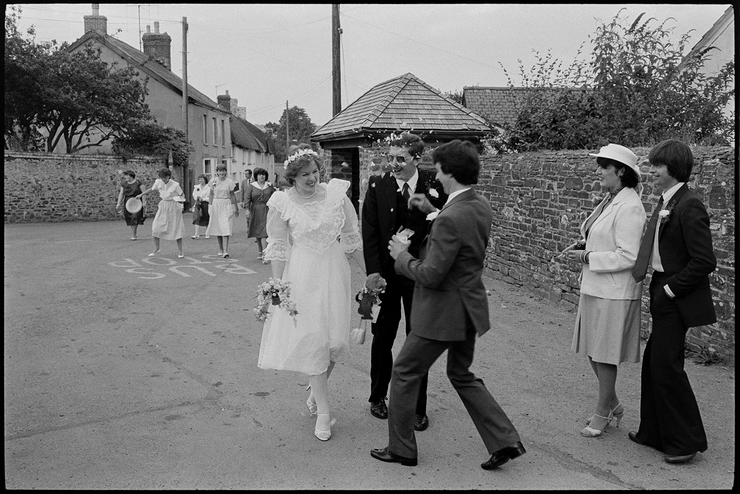 Wedding, groups, bride and groom leaving church, chatting to guests. 
[A man throwing confetti over Cynthia Palmer and her groom after their wedding in Dolton. They are walking along a street past a bus shelter. Other wedding guests are following them and watching.]