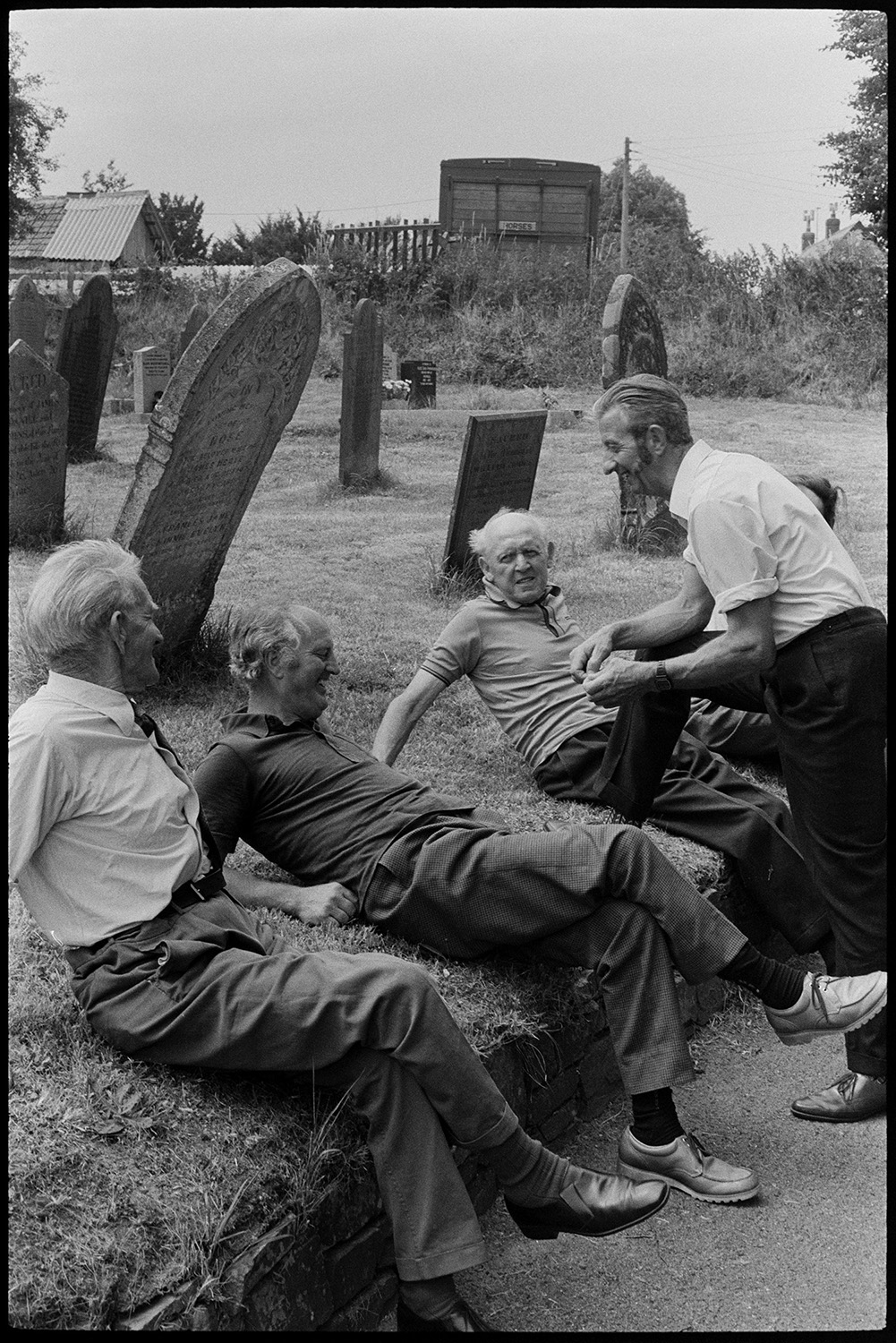 Bellringers resting after wedding, guests chatting and taking photographs. 
[Five men resting on the grassy bank in Dolton churchyard after bell ringing for Cynthia Palmer's wedding. Gravestones can be seen in the background.]