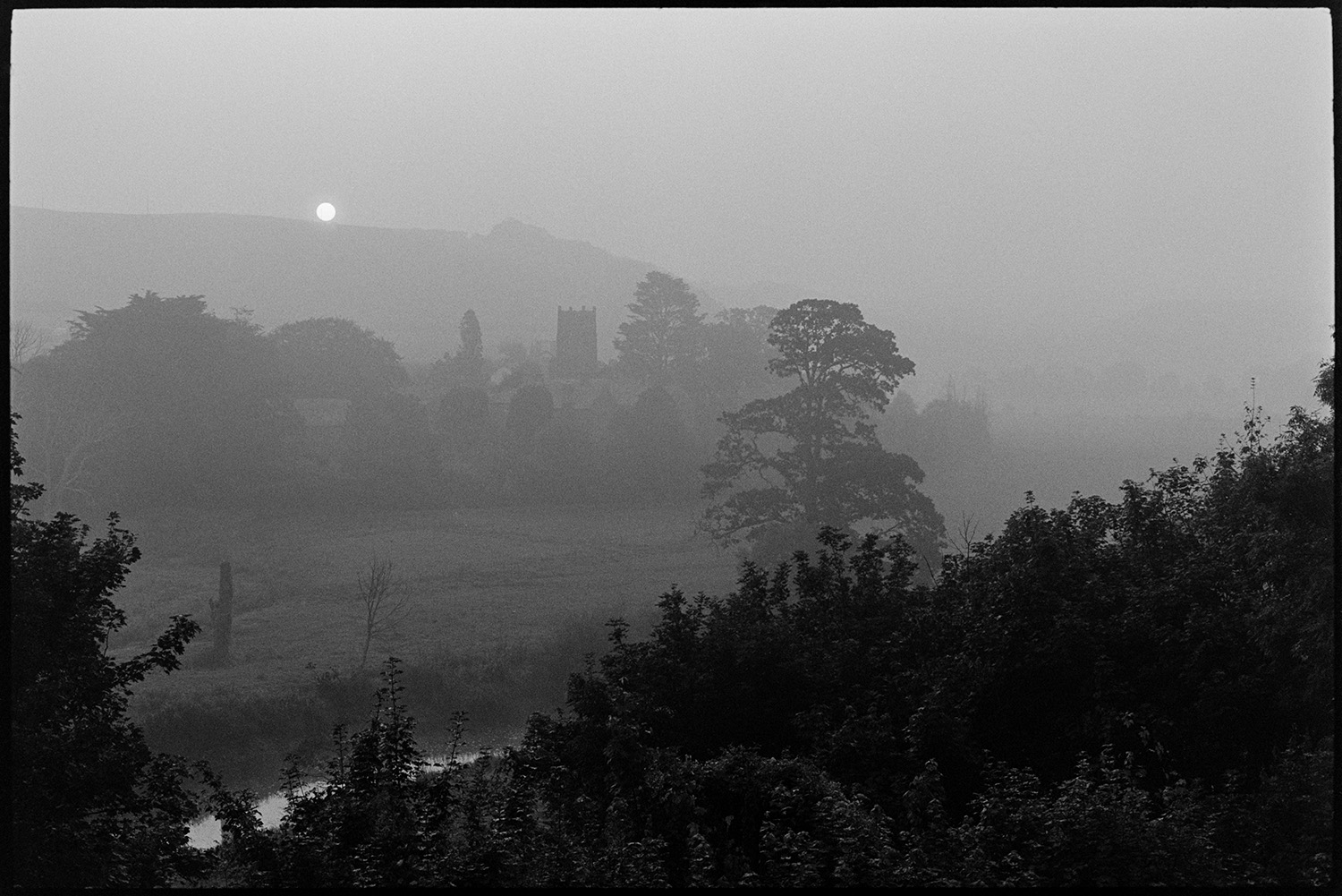 Sun rising over river and church. 
[The sun rising over a landscape at Weare Giffard. The church, surrounded by trees, is visible and the River Torridge can be seen in the foreground.]