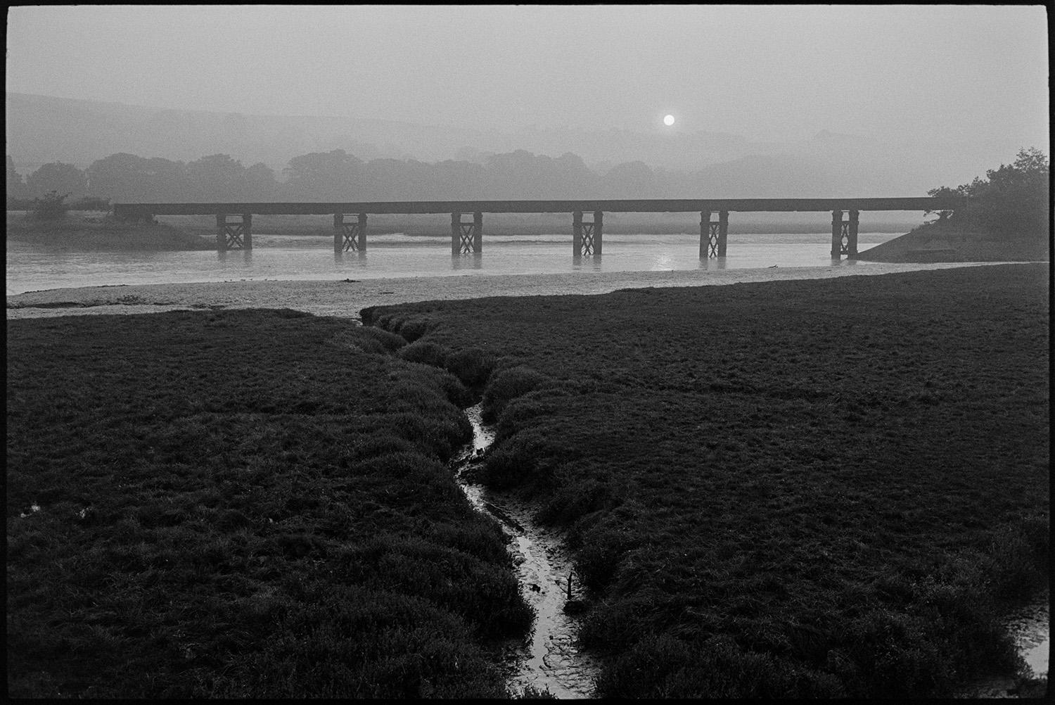 Sun rising over railway bridge over river. 
[The sun rising over a landscape at Pillmouth, Landcross with the railway bridge crossing the River Torridge. Marshland is visible in the foreground.]