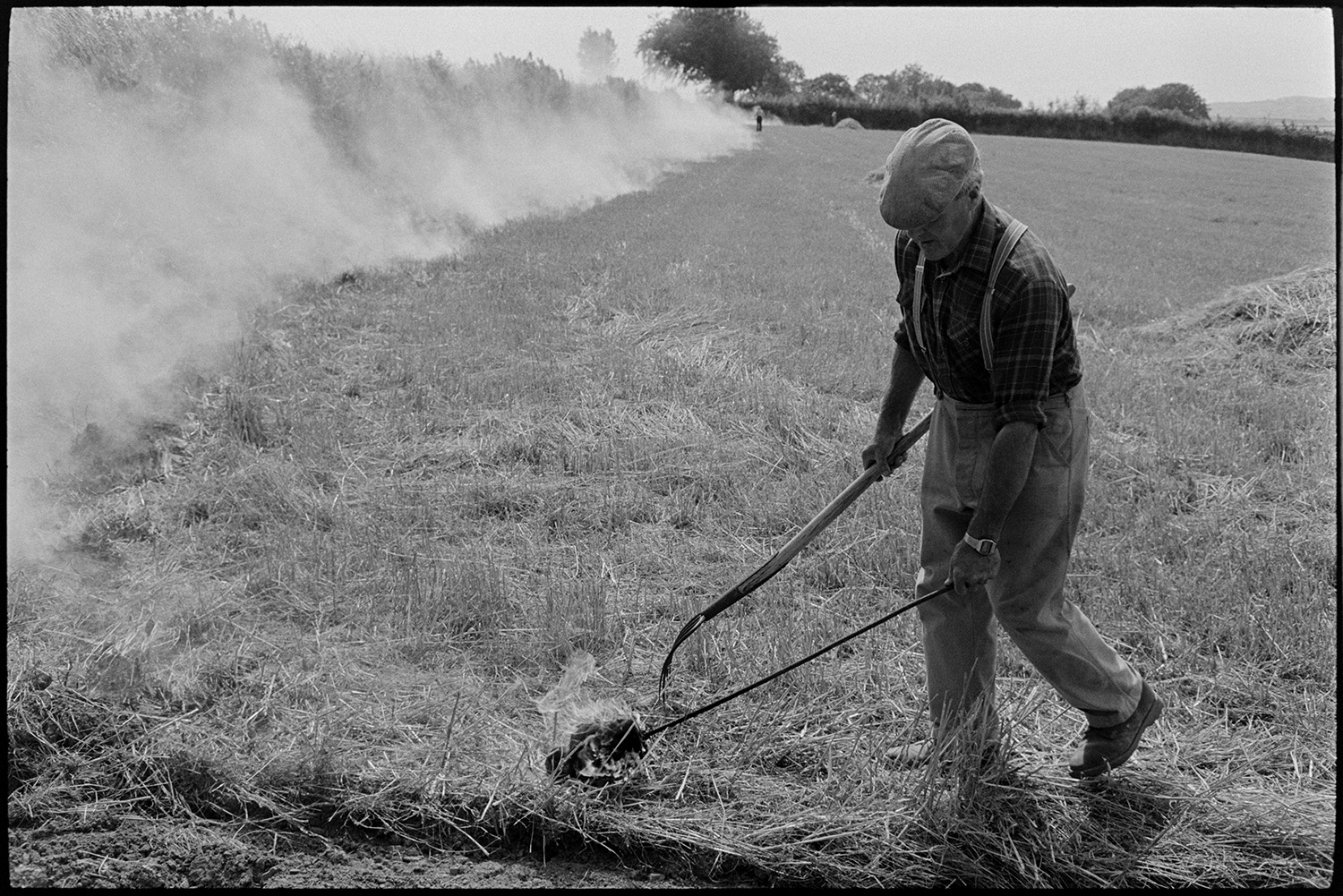 Farmer burning stubble. 
[Alf Pugsley burning crop stubble in a field at Lower Langham, Dolton. He is using a flaming torch and a fork. Smoke is rising from the burnt areas in the background.]