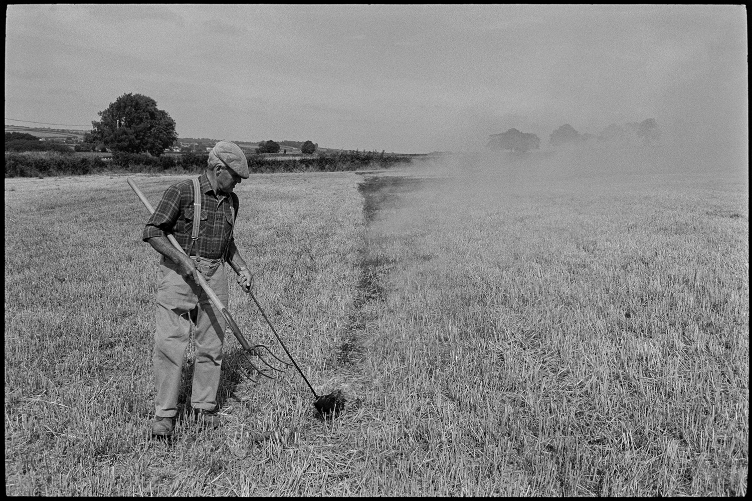 Farmer burning stubble. 
[Alf Pugsley burning crop stubble in a field at Lower Langham, Dolton. He is using a flaming torch and a fork. Smoke is rising from the area he has already burnt.]