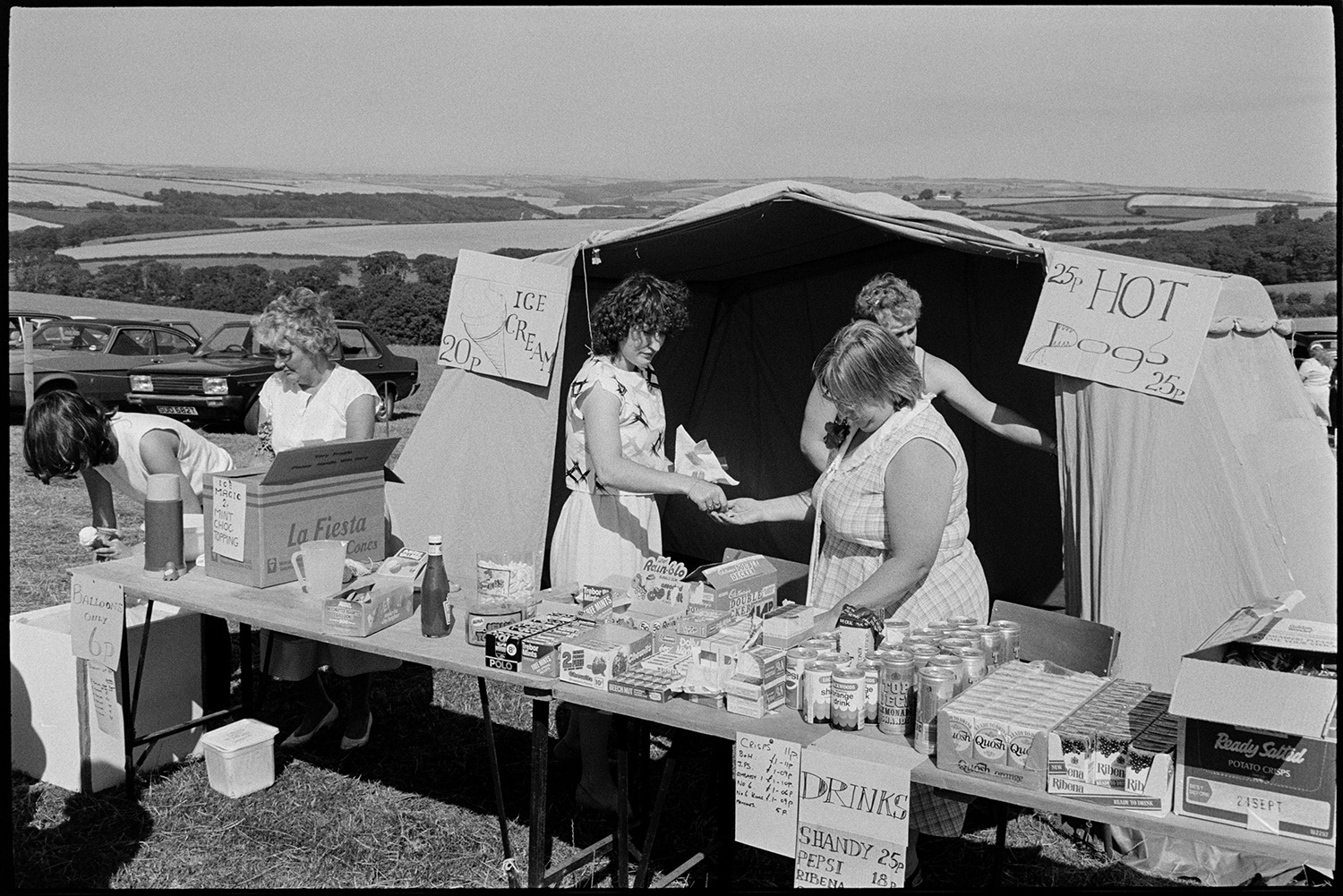 Fete, produce, stalls, machinery, flowers. 
[Women running a refreshment stall and tent at a fete at either Monkleigh or Frithelstock. They are selling drinks, sweets, ice creams and hot dogs.]