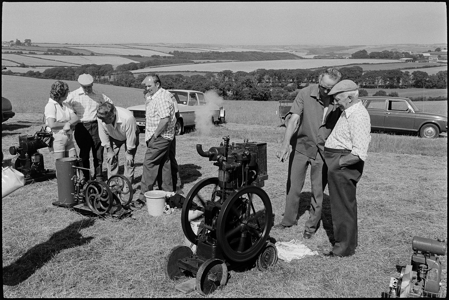 Fete, produce, stalls, machinery, flowers. 
[Men and a woman looking at machinery or traction engines in a field at a fete at either Monkleigh or Frithelstock. Cars are parked in the background in front of a landscape of trees and fields.]
