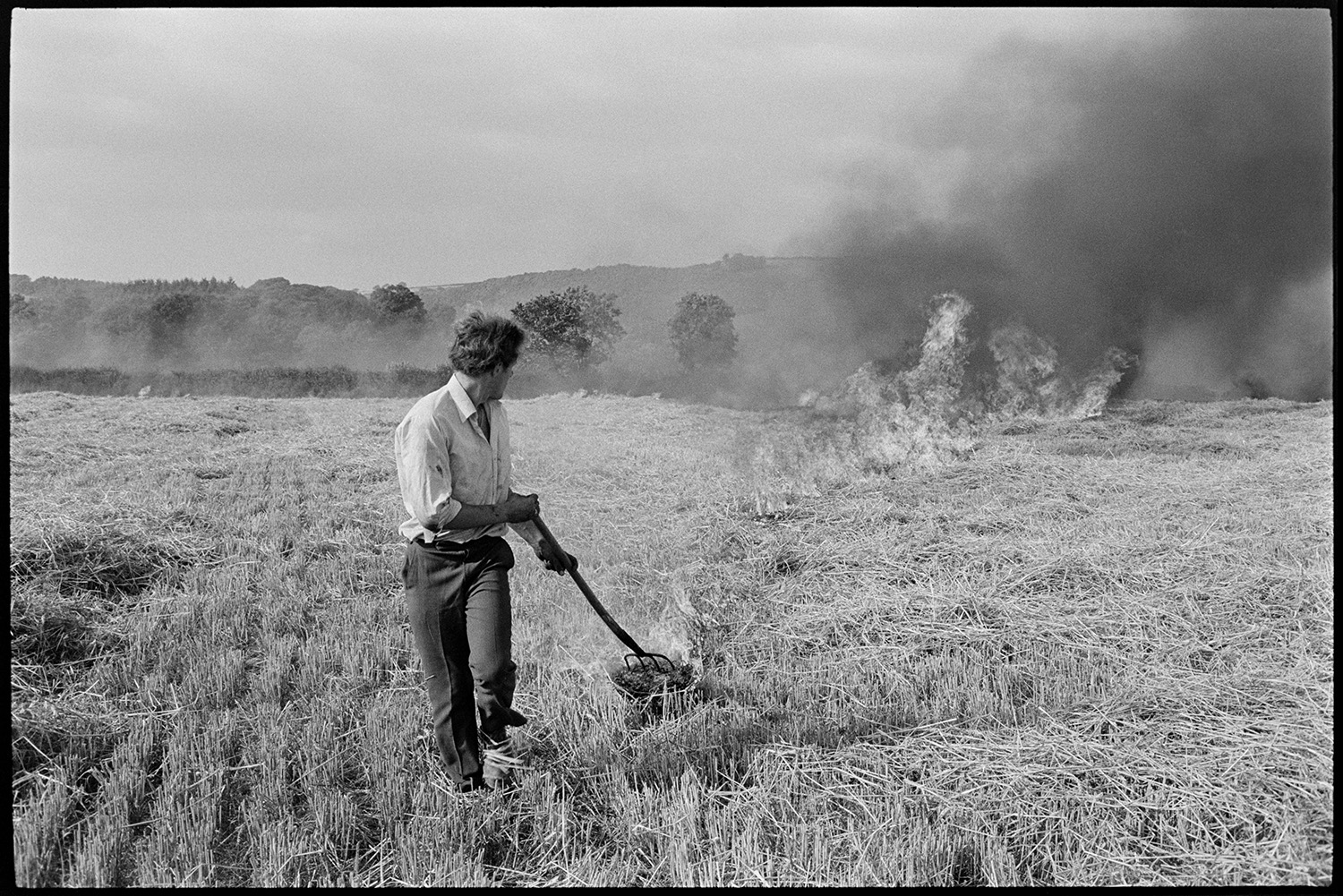Farmer burning stubble. 
[A man burning crop stubble in a field at Greatwood, Merton. He is using a fork to light the stubble. Black smoke is rising from the stubble already alight.]
