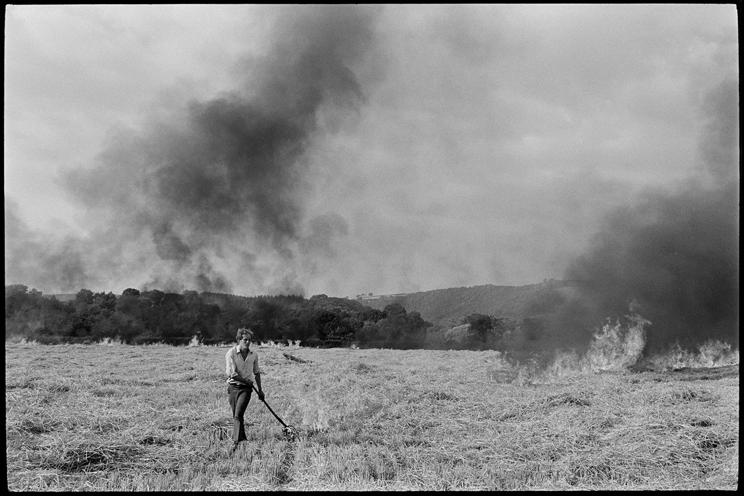 Farmer burning stubble. 
[A man burning crop stubble in a field at Greatwood, Merton. He is using a fork to light the stubble. Black smoke is rising from the stubble already alight and woodland can be seen in the background.]