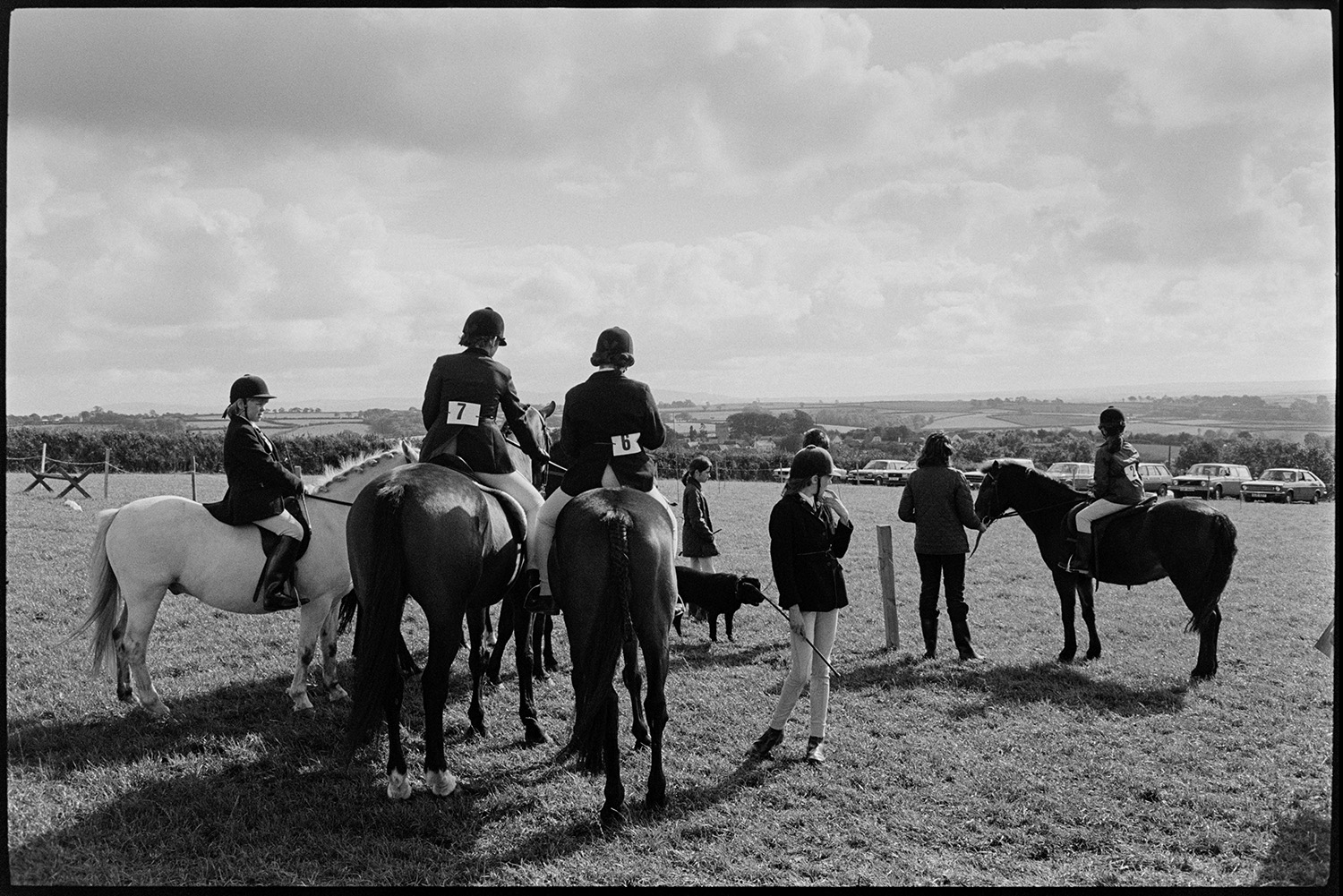 Horses and riders at gymkhana. 
[Mounted horse riders waiting to compete at Dolton Gymkhana. Some of the competitors are children. Parked cars can be seen in the field in the background.]