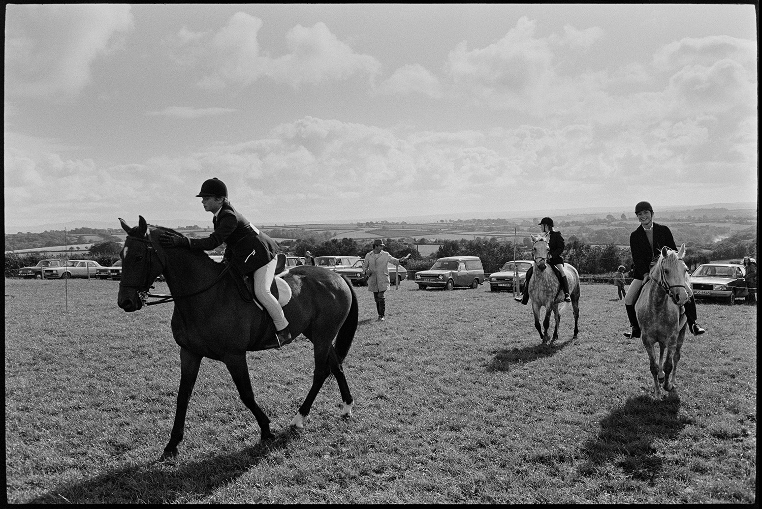 Horses and riders at gymkhana. 
[Three mounted horse riders at Dolton Gymkhana. Parked cars can be seen in the field in the background.]