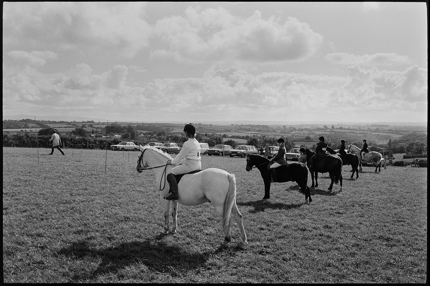 Horses and riders at gymkhana. 
[Mounted horse riders lined up ready to compete in an event at Dolton Gymkhana. Parked cars can be seen in the field in the background.]