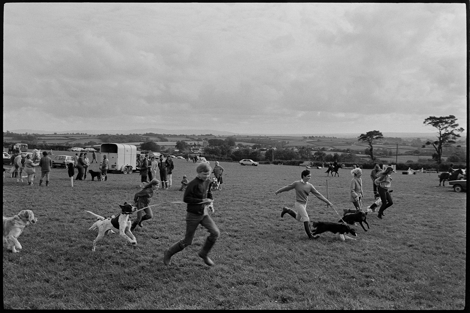 Dogs and owners at dog show, being judged.
[Dog owners running with dogs on leads at a dog show in a field at Dolton Gymkhana. Men, women and children are watching. In the background horses are being ridden across the field, and horse boxes, parked cars and a view of the surrounding countryside are visible.]
