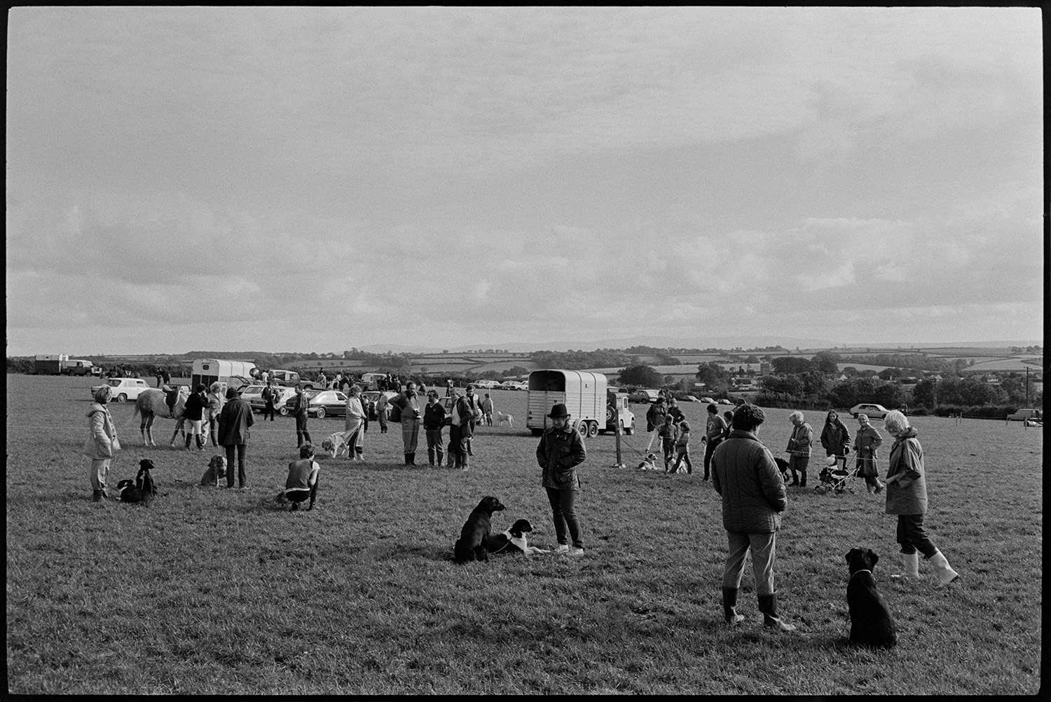 Dogs and owners at dog show, being judged.
[Dogs sat by their owners and being judged in a dog show at Dolton Gymkhana. The judging is being watched by men, women and children. A saddled horse, horse boxes and cars are visible in the background, and a view of surrounding countryside.]