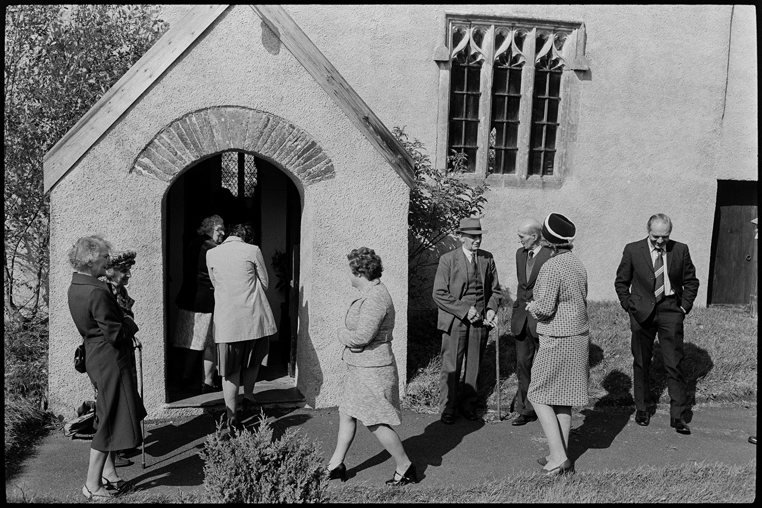 People going into church, clothes, Sunday best dresses and suits, church porch and window.
[Men and women entering the Church of St. Peter, Dowland for the Harvest Festival. They are wearing suits, dresses and hats. In addition to the church porch entrance, the detail of the window to the right of the porch can be seen.]