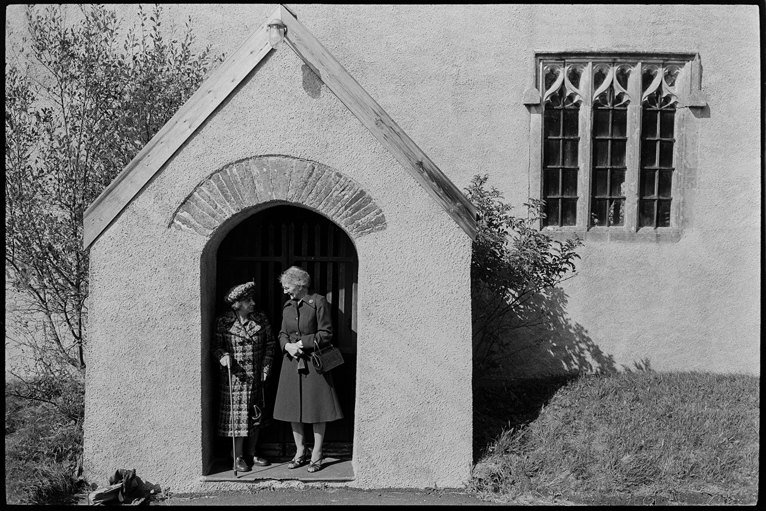 People going into church, clothes, Sunday best dresses and suits, church porch and window. 
[Two women dressed in smart coats and hats, one with a walking stick, standing and chatting in the porch of the Church of St. Peter, Dowland, after attending the Harvest Festival. Details of the window to the right of the porch can be seen.]