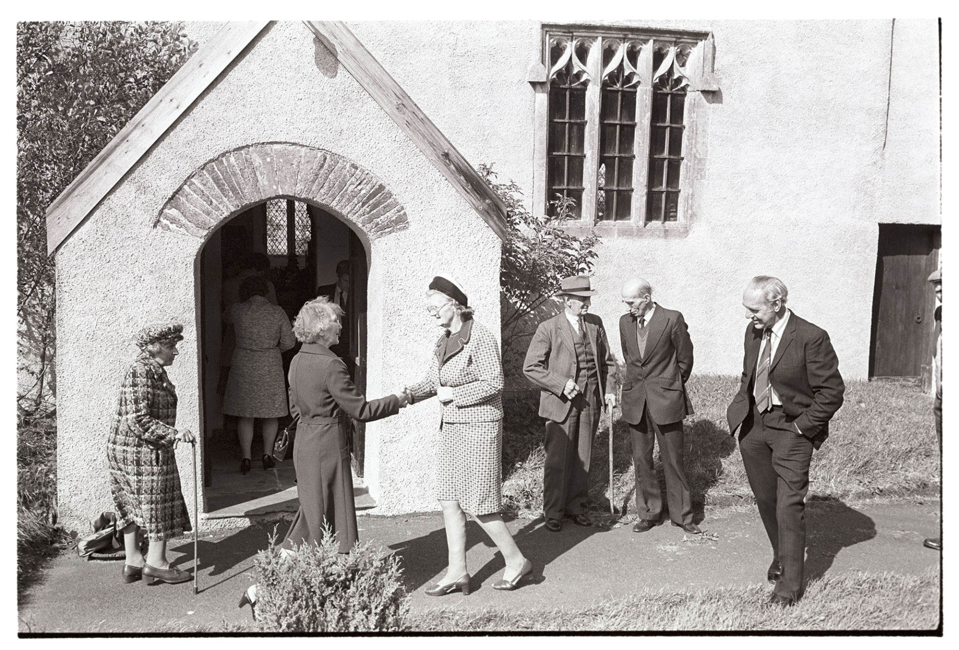 People going into church, clothes, Sunday best dresses and suits, church porch and window.
[A group of men and women outside the porch of Dowland Church, about to attend the Harvest Festival service.  Two women are shaking hands.]