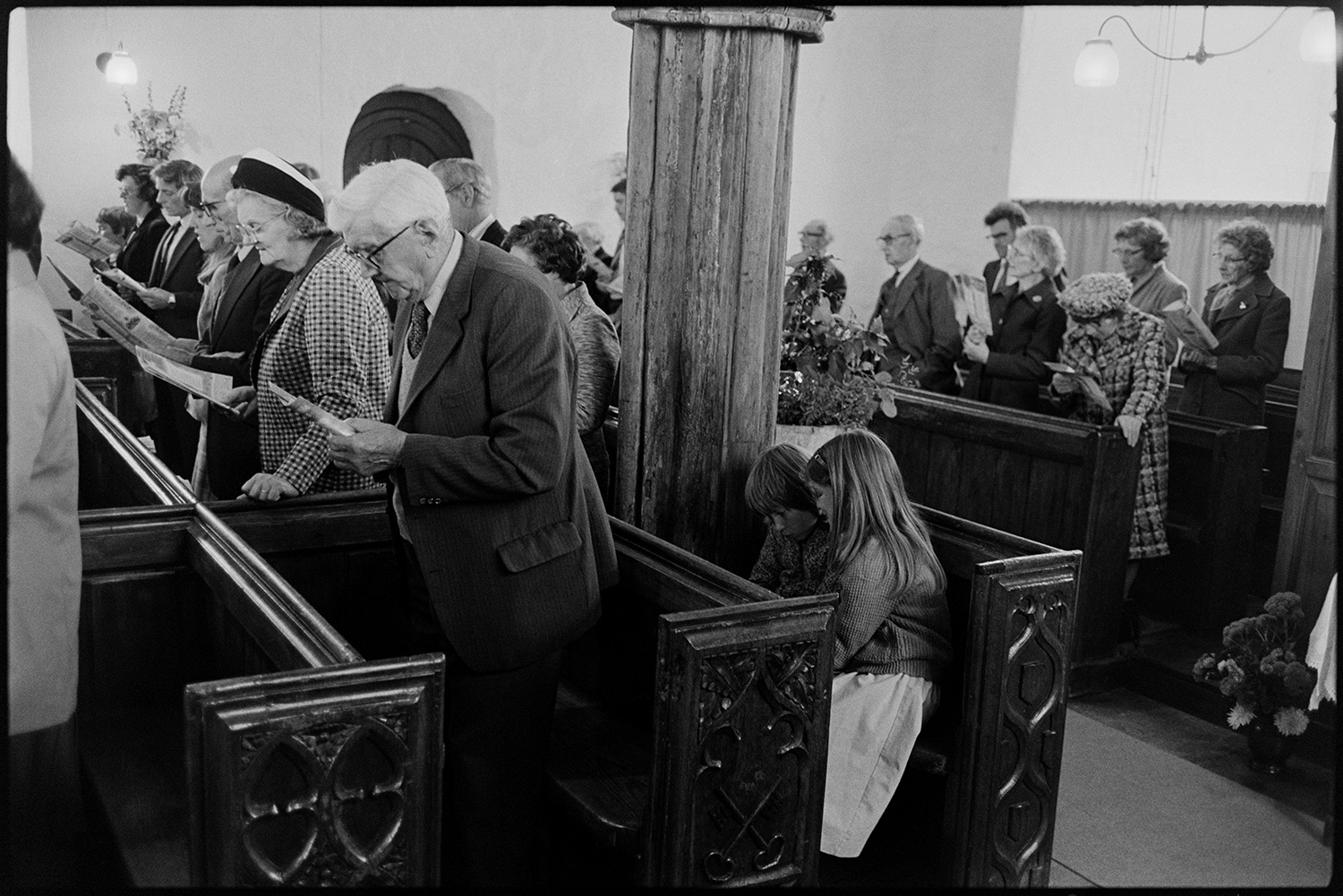 People in church, vicar preaching sermon, pulpit.
[Men, women and children dressed in suits and smart clothes standing in wooden pews in the Church of St. Peter, Dowland, singing hymns at the Harvest Festival. Some of the detailed wood carving on the end of the pews can be seen.]