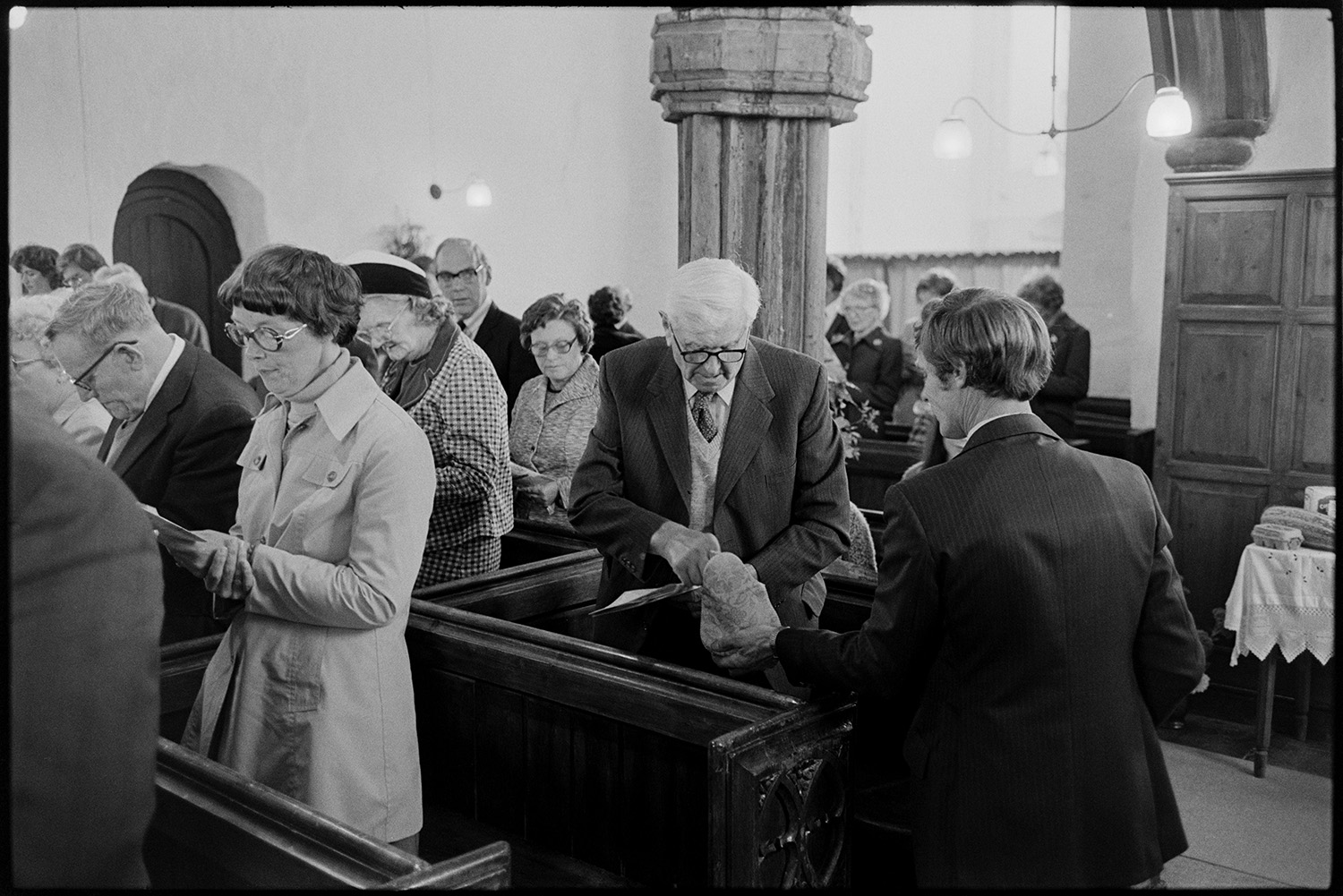 People in church singing hymns.
[Men and women standing in wooden pews in the Church of St. Peter, Dowland, singing hymns at the Harvest Festival. One man in the congregation is putting money into a collection bag being held out by another man. One of the church pillars and a wood carving on the end of a pew can be seen.]