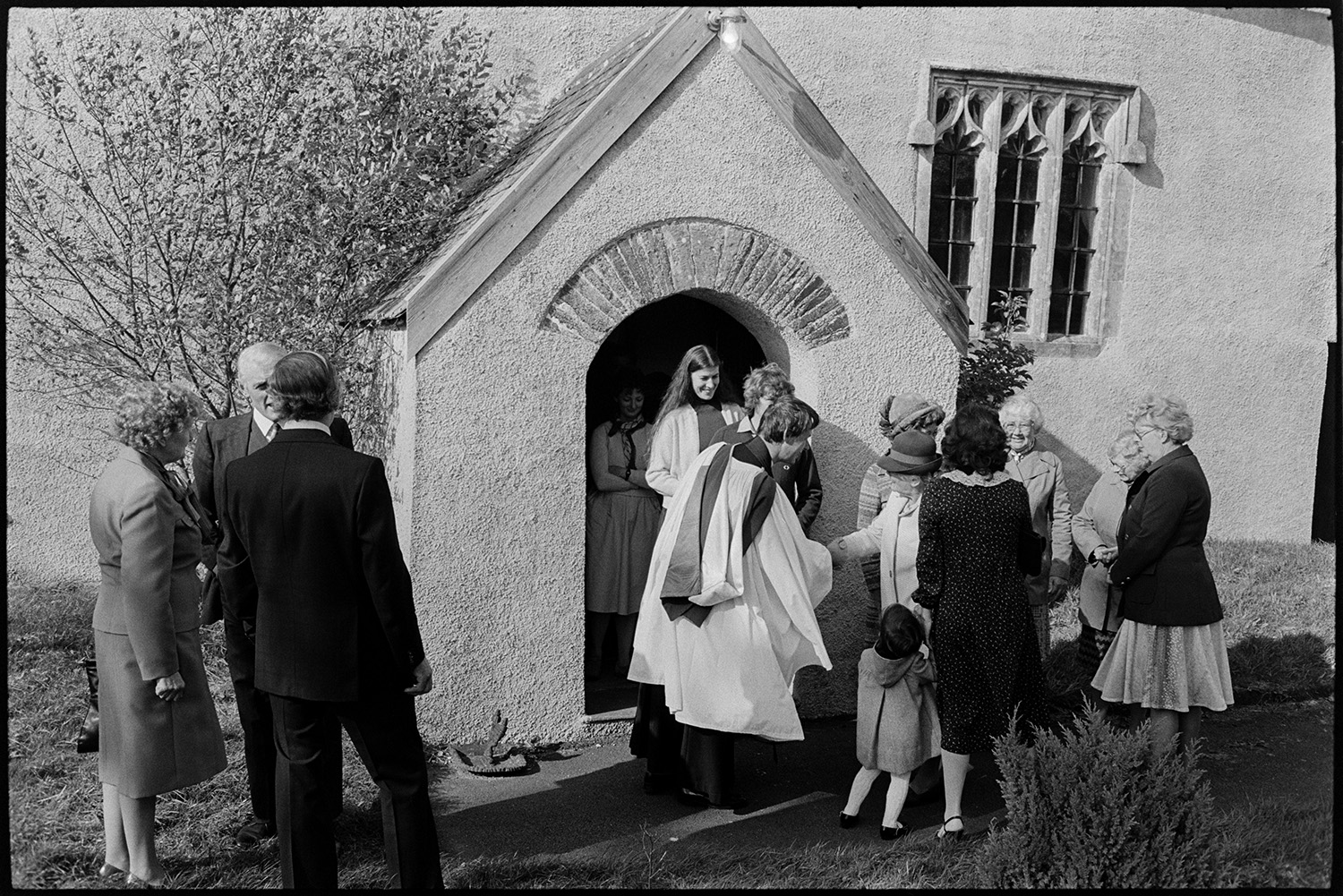 People coming out of church vicar saying hello and goodbye. Clothes.
[Reverend Jeremy Bell shaking hands and talking to members of the congregation, in front of the porch of the Church of St. Peter, Dowland, as they leave the church after the Harvest Festival. The detail of the window to the right of the porch can be seen.]