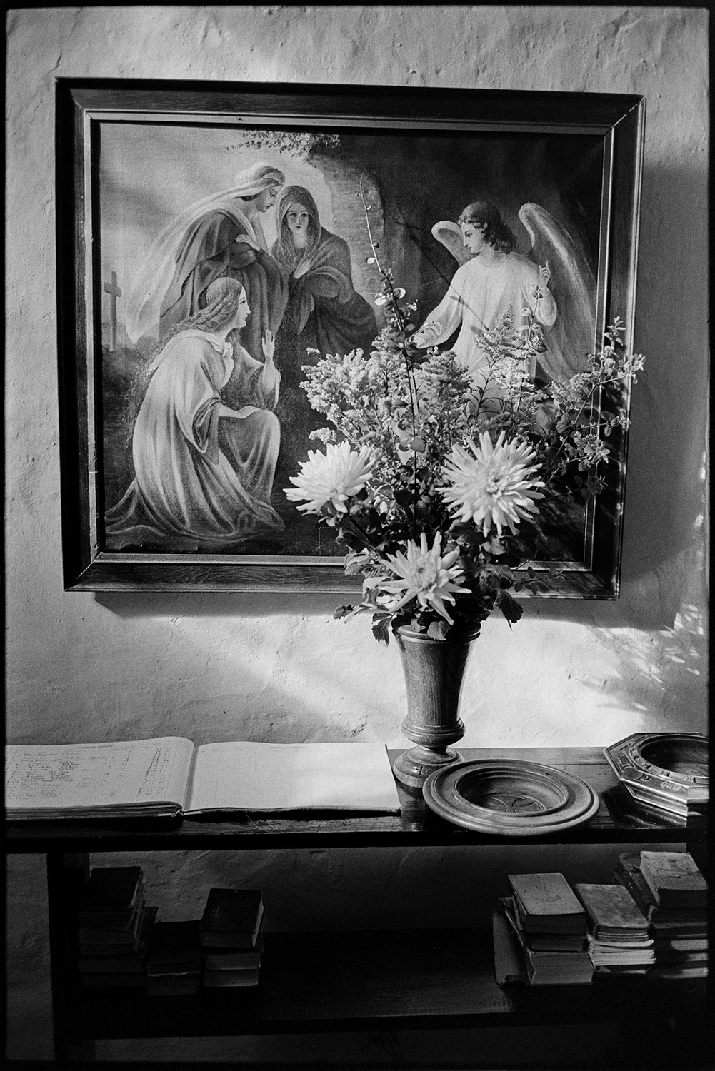 People coming out of church vicar saying hello and goodbye. Clothes.
[Interior of the Church of St. Peter, Dowland, showing a religious painting mounted on wall behind a vase of flowers standing on a bookshelf, with an open ledger book, offertory dishes and books. The scene is lit by sunlight.]