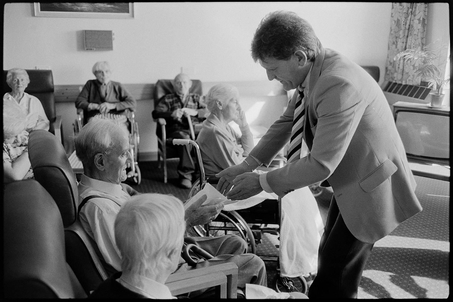 TV presenter at hospital event cutting cake.
[Ian Stirling, a TV presenter, handing a piece of cake to an elderly patient at an event in Barnstaple General Hospital. Other patients are sitting in chairs around the room.]