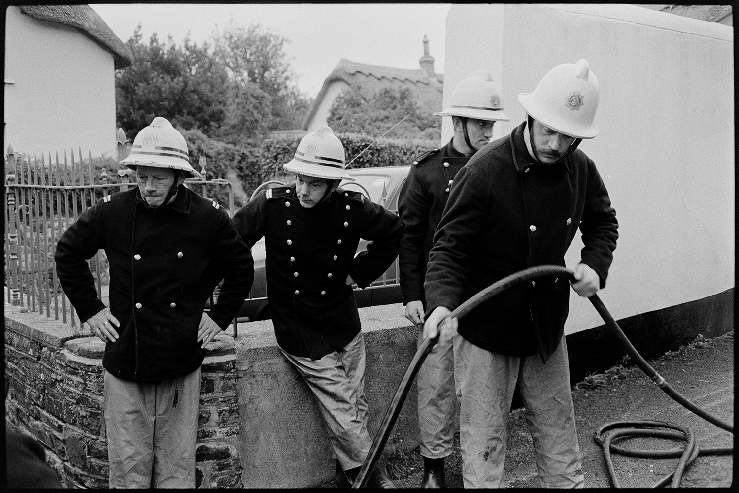 Firemen in uniform at fire. Chatting afterwards, helmets.
[Four firemen from the Hatherleigh Fire Brigade working on an incident in Fore Street, Dolton. One fireman is holding a hose. A house with a thatched roof is visible in the background.]