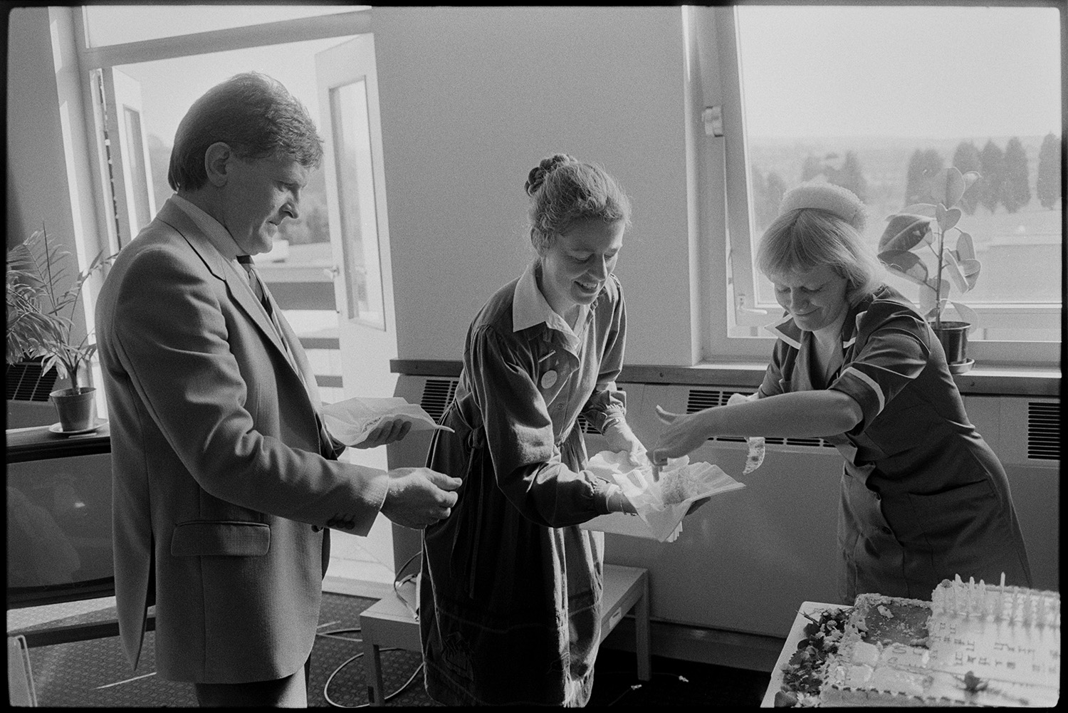 TV presenter at hospital event cutting cake.
[Ian Stirling, a TV presenter, and Diana Johnson collecting pieces of cake from a nurse to give to patients at an event in Barnstaple General Hospital.]