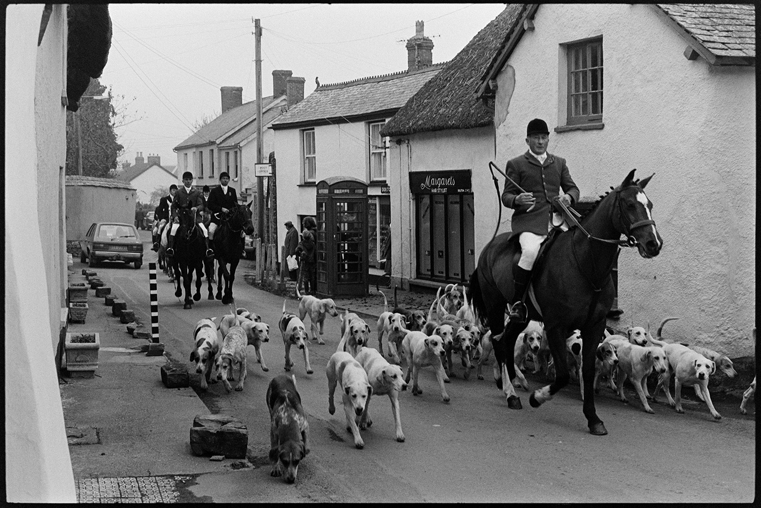 Hunt meet, horses and riders in village in front of pub. 
[Horse rider and hounds setting off on a hunt along Fore Street, Dolton, after their meet outside the Royal Oak pub. They are passing Margaret's hair salon and the telephone box.]