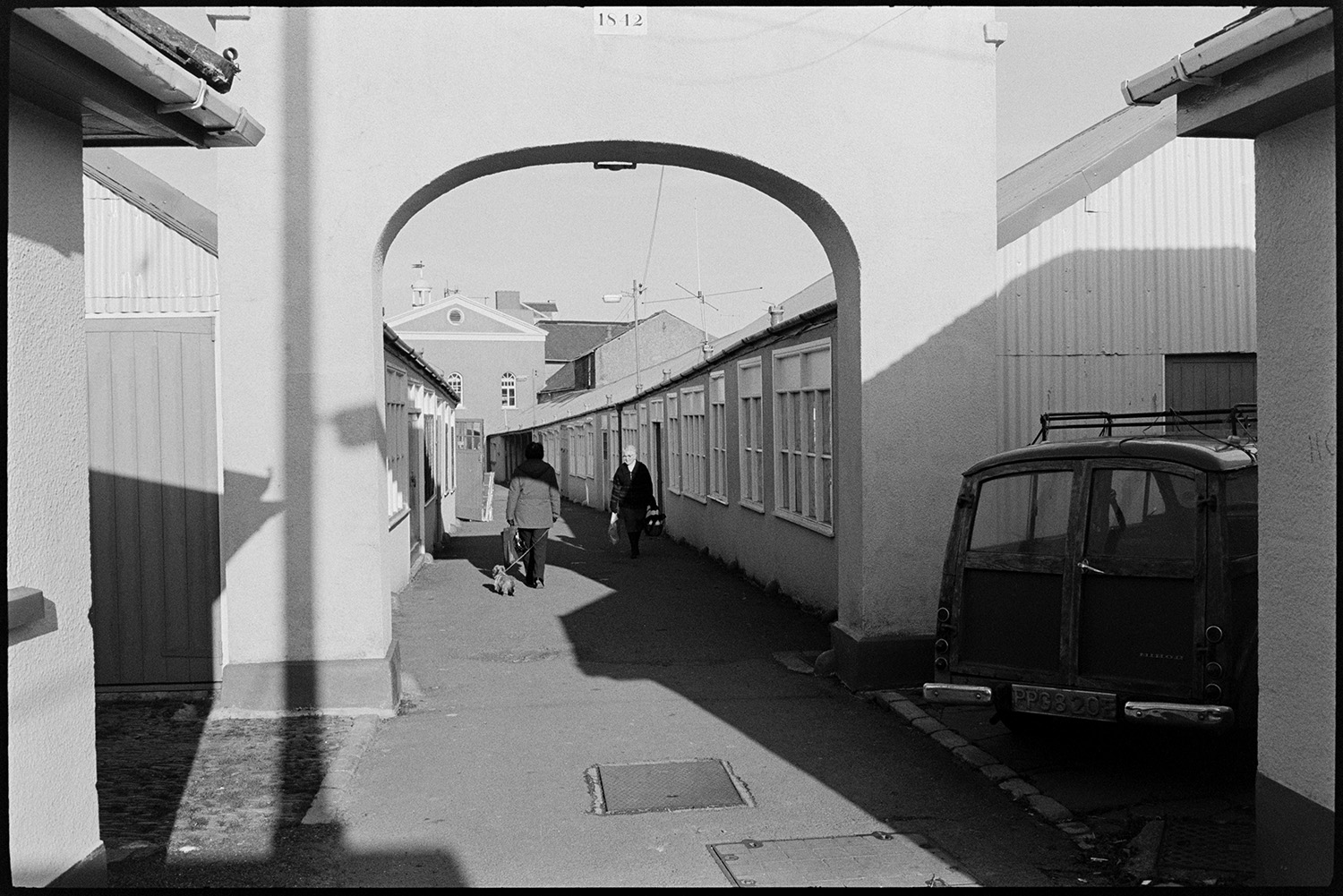 View through arch to market. 
[A van parked by the archway to Torrington Market. Two people are walking past the shops in the market. One of them is walking a dog.]