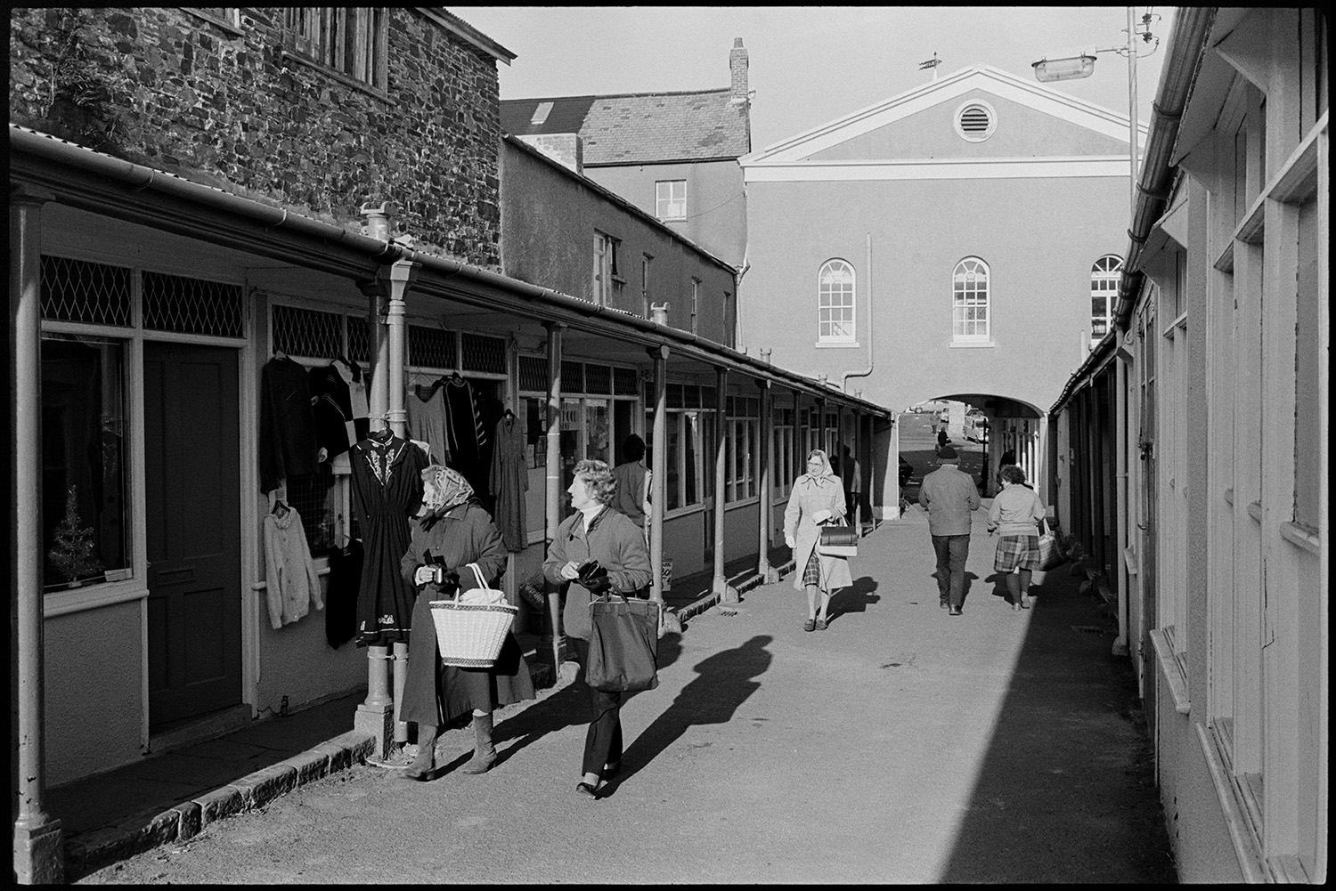 Street scenes with decaying front of antique shop, market.
[People shopping in Torrington Pannier Market. Two women are looking at clothes hung up outside one of the shop fronts.]