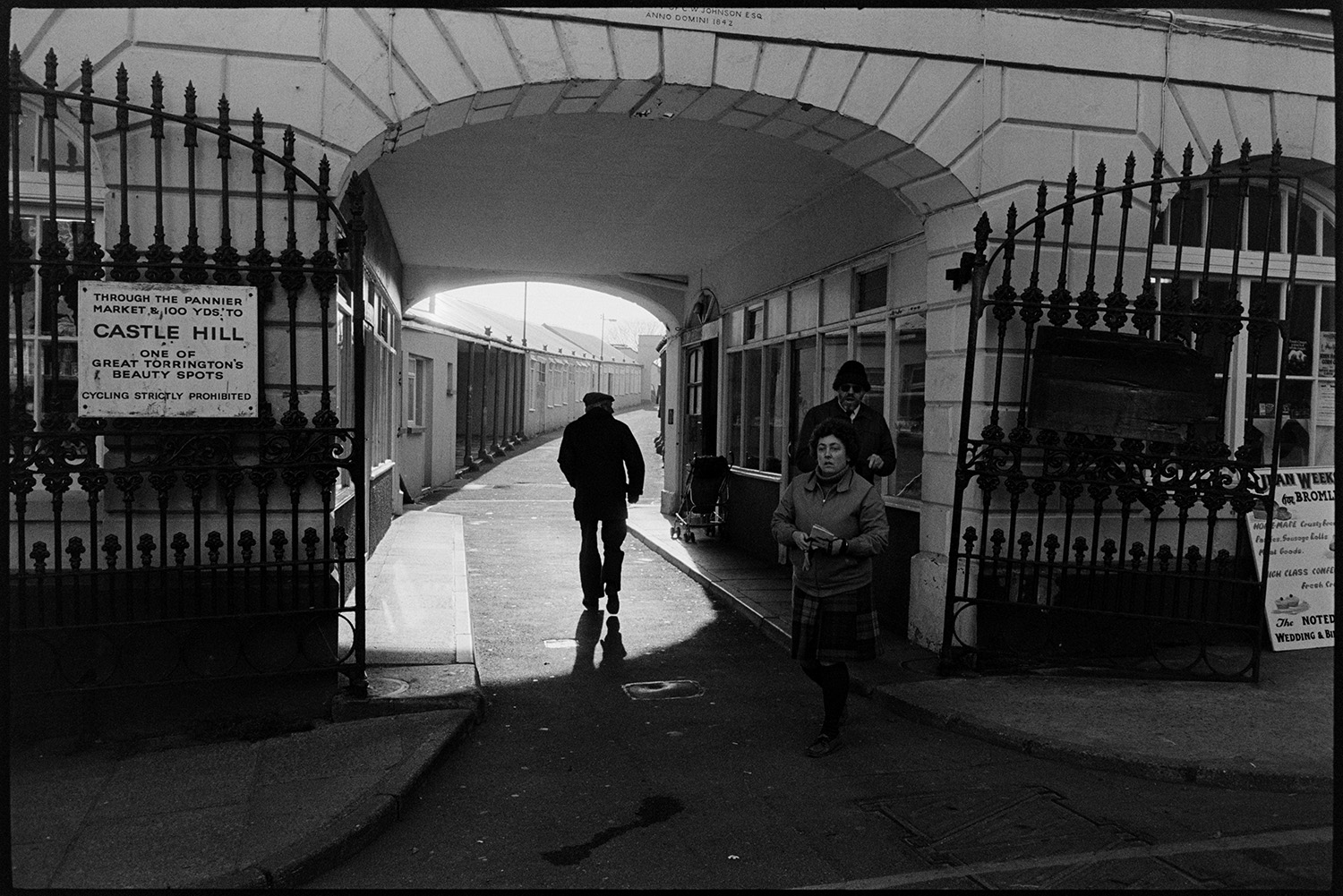 Street scenes with decaying front of antique shop, market.
[People walking under the archway entrance of Torrington Pannier Market. A sing on the open railings to the market is advertising Castle Hill beauty spot.]