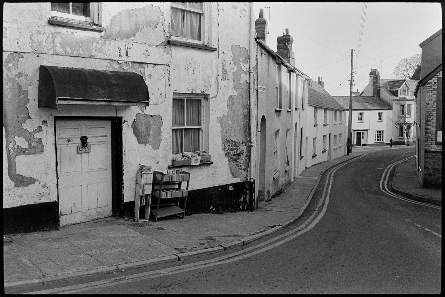 Street scenes with decaying front of antique shop, market.
[A view of Castle Street, Torrington with a crumbling house or antique shop with books for sale outside displayed on bookshelves and the windowsill.  An open sign is on the door knocker and an awning is above the doorway.]