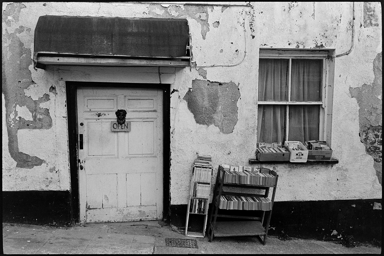 Street scenes with decaying front of antique shop, market.
[The front of a house or antique shop with crumbling plaster with books for sale outside displayed on bookshelves and the windowsill, in Castle Street, Torrington  An open sign is on the door knocker and an awning is above the doorway.]