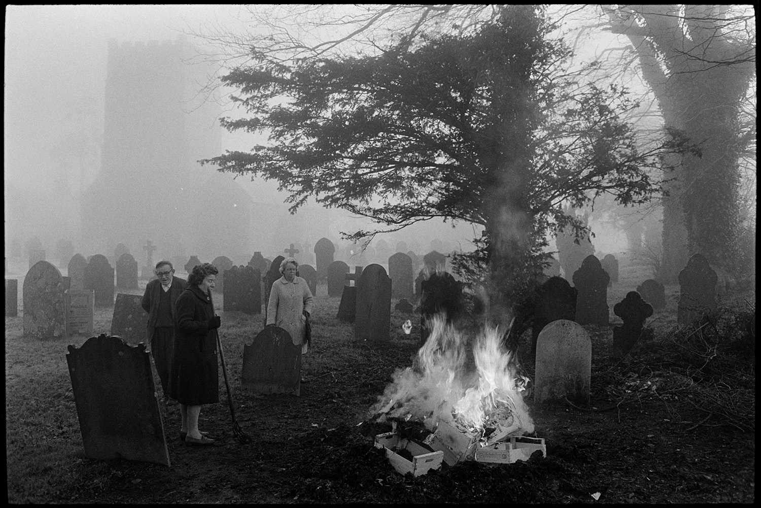People burning rubbish from village stores on graveyard bonfire, misty church behind.
[Members of the Friend family standing by a bonfire in the misty churchyard at Dolton Church. They are burning rubbish from the village stores, including boxes. Gravestones and the church can be seen through the mist in the background.]