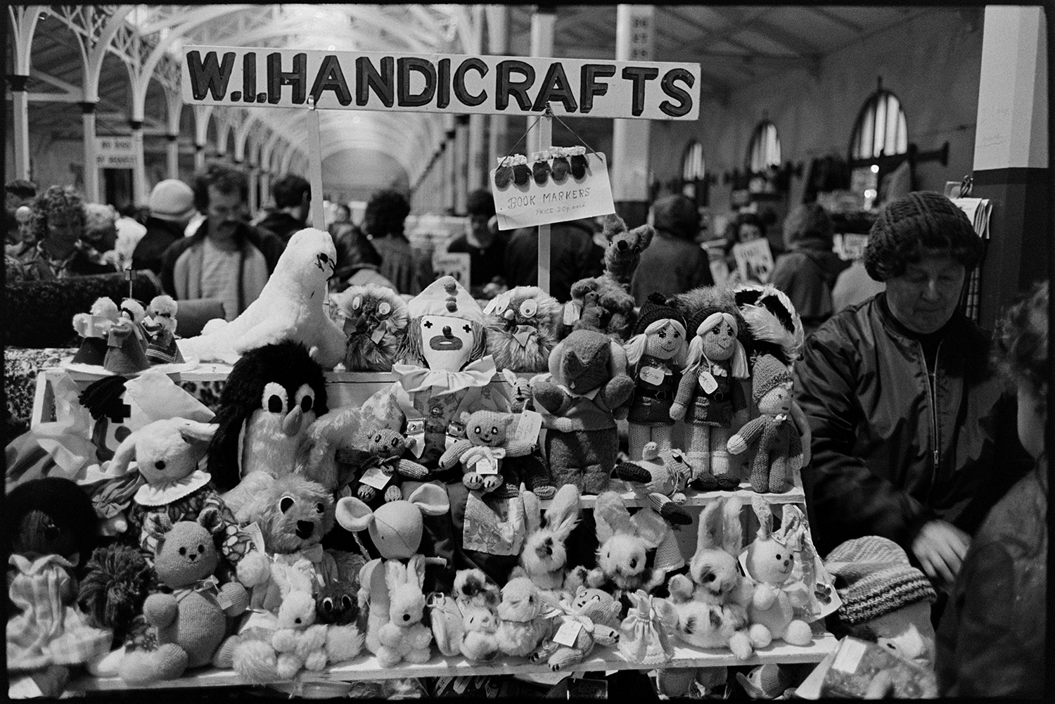 WI Handicrafts stall in pannier market. Stuffed toys.
[Handmade soft toys, including teddy bears, rabbits and dolls, on the Women's Institute Handicrafts stall in Barnstaple Pannier Market.  People shopping and stall holders are visible in the background.]