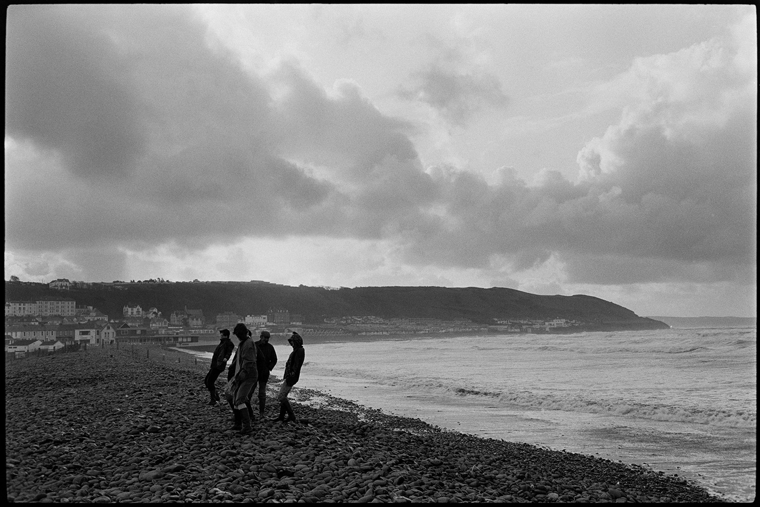 People walking beside stormy sea, leaning on the wind.
[Lord Clinton and friends standing on Westward Ho! pebble ridge, leaning into the wind. Views of the beach and coastal buildings are visible in the background.]