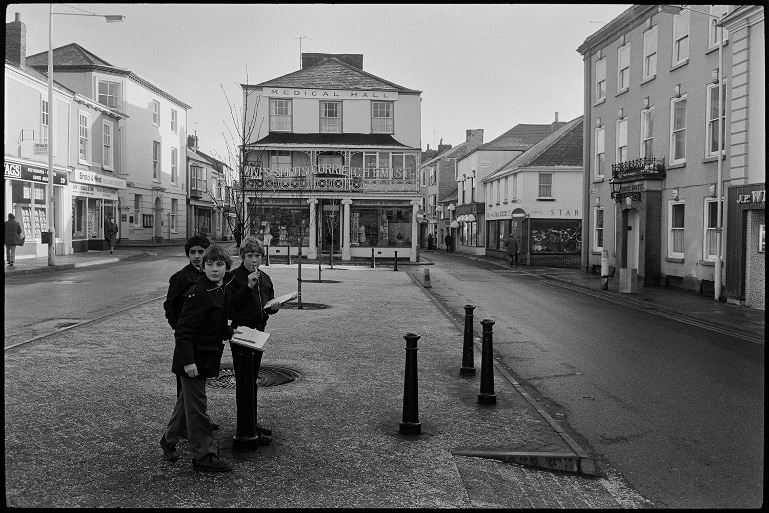 Schoolchildren doing project in town, chemist's shop behind.
[Street scene with three school children in the square in South Molton for a school project.  The George Hotel is visible on the right and the Medical Hall or Currie's Chemist can be seen in the background.]