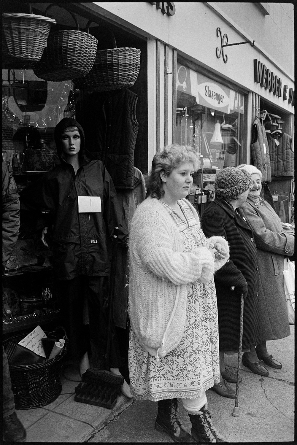 Woman waiting outside shop.
[Women standing outside Webber & Son shop in South Molton. Two women chatting. Baskets and a mannequin modelling clothes are outside the shopfront.]