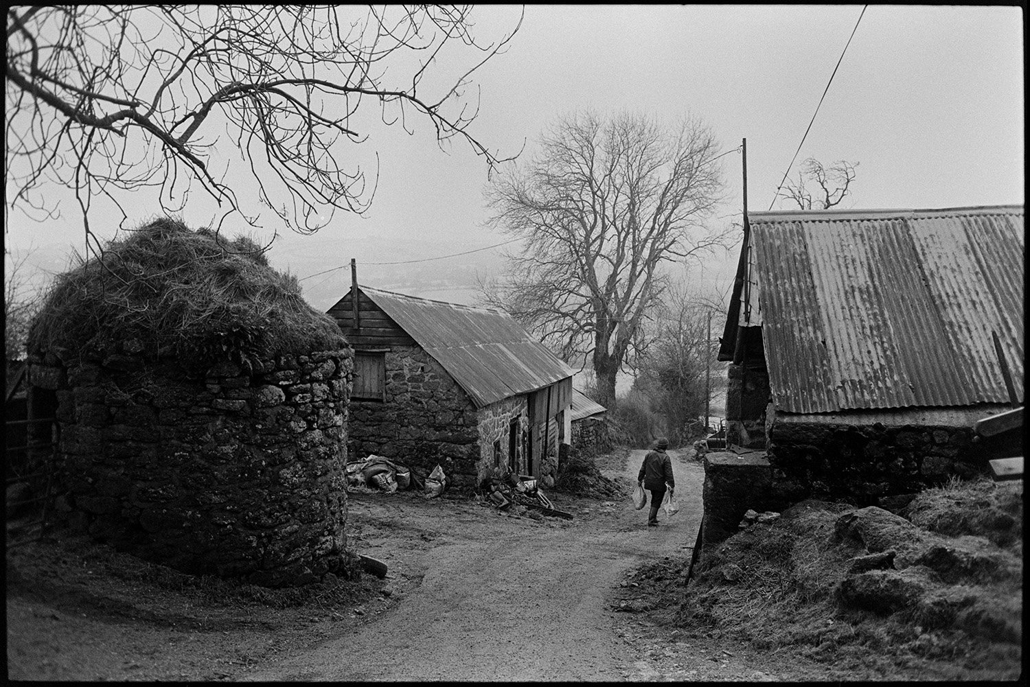 Snow, farmers feeding cattle on moor, photographer taking pictures.
[A farmer carrying sacks down a farm lane between two stone barns with corrugated iron roofs near Chagford, Dartmoor.]