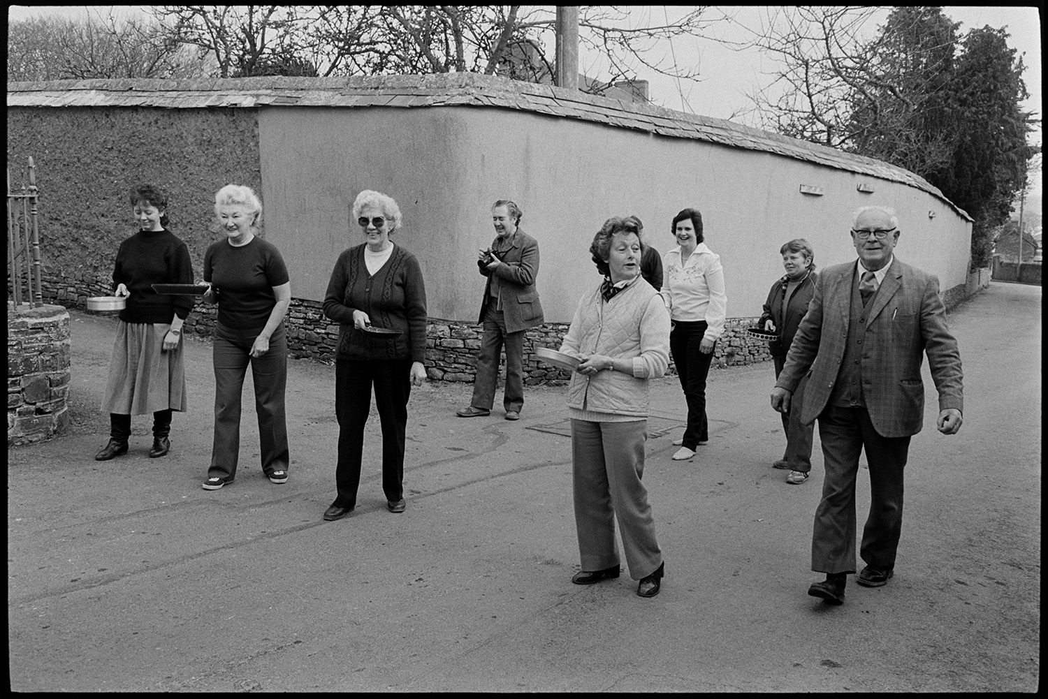 Children and parents at Pancake Race in village street.
[Women lined up by a wall in Fore Street, Dolton at the start line of a pancake race. A man in the background has a camera to photograph the race.]