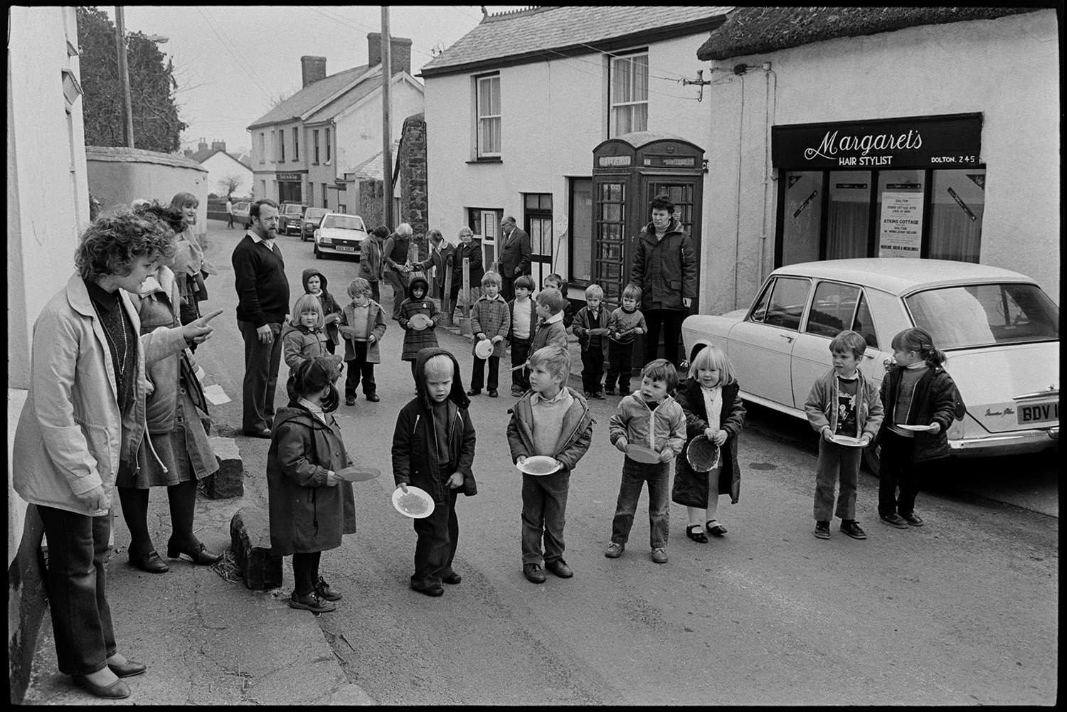Children and parents at Pancake Race in village street.
[Children lining up outside Margaret's hair salon and telephone box in Fore Street, Dolton for a pancake race. Men and women are watching.]