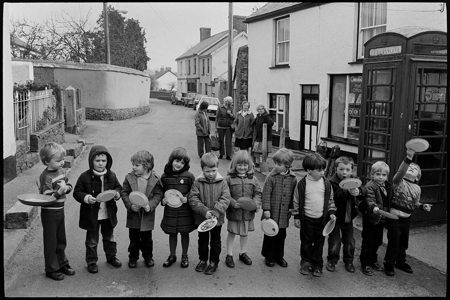 Children and parents at Pancake Race in village street.
[Children lining up by the telephone box in Fore Street, Dolton for a pancake race. A group of women are chatting in the background.]