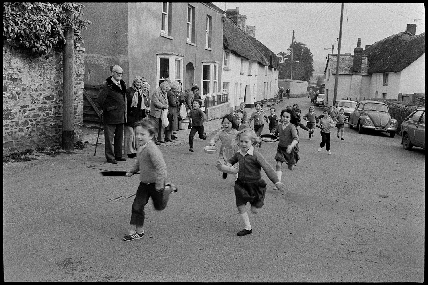 Children and parents at Pancake Race in village street.
[Children running along Fore Street, Dolton in a pancake race. A line of adults are watching from the side of the street.]