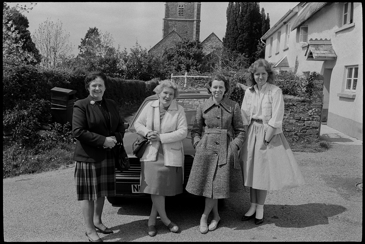Club Day, people waiting before parade, Silver Band and club members setting off, drinkers.
[Four women standing in front of a car parked outside a house near Iddesleigh church on Iddesleigh Club Day.]