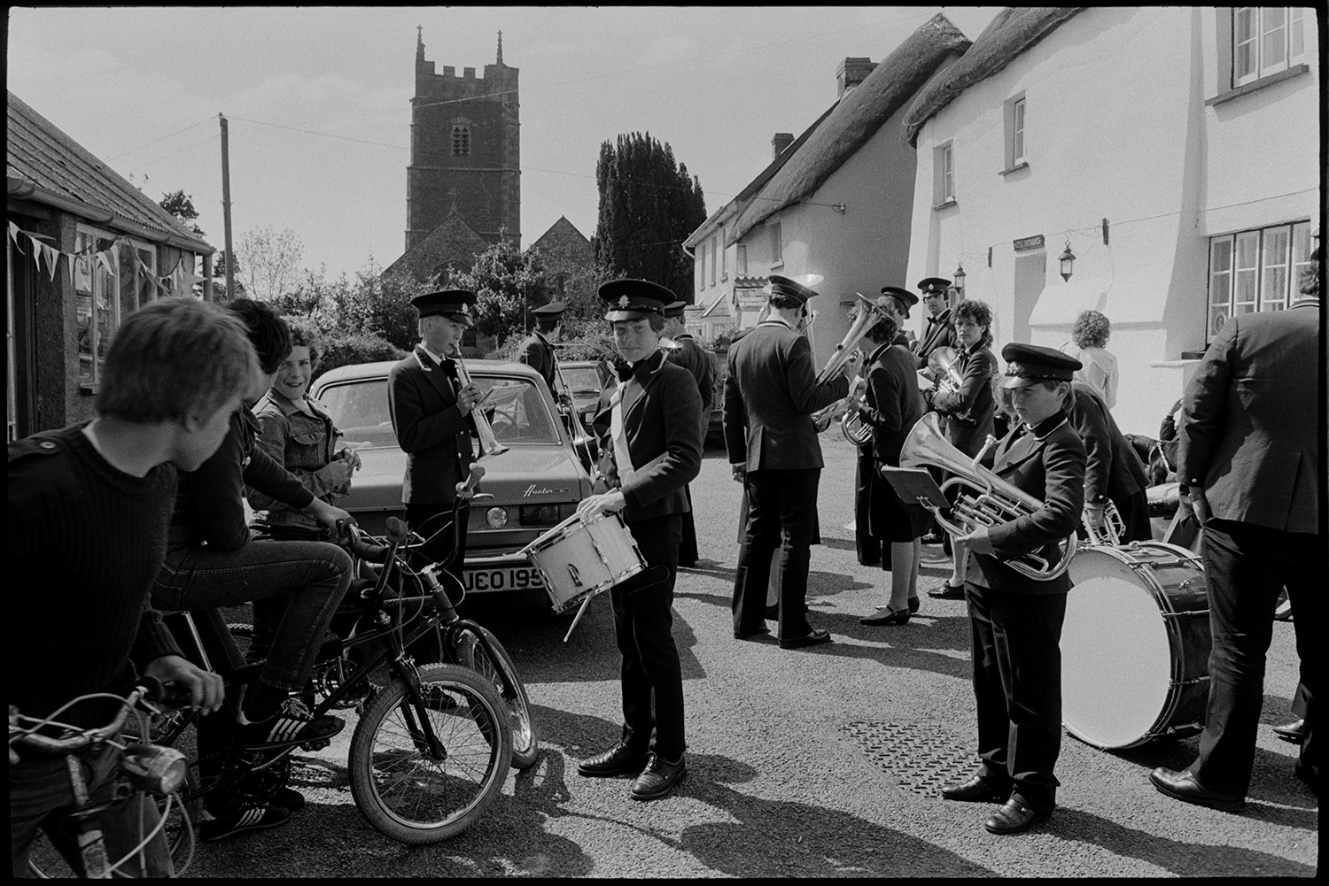 Club Day, people waiting before parade, drinking, playing cards. Boys on bicycles looking on.
[Members of the Hatherleigh Silver Band and boys on bicycles gathered outside the Duke of York Inn on Iddesleigh Club Day, before the Club Day parade. A Hillman Hunter car is parked in the street and Iddesleigh Church can be seen in the background.]