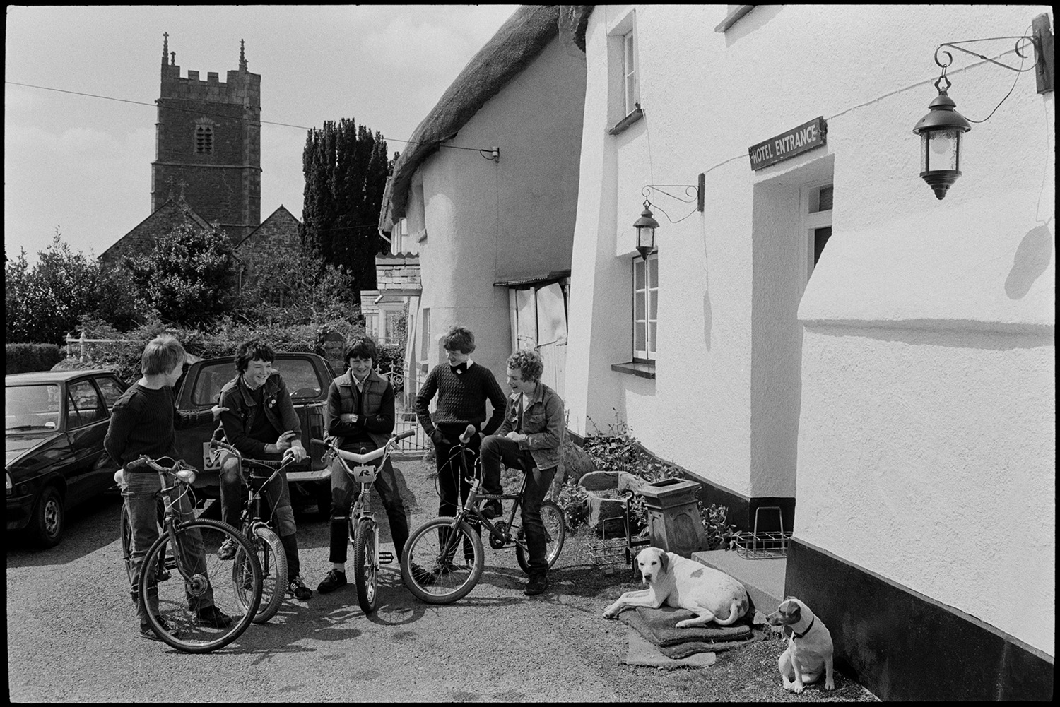Club Day, people waiting before parade, drinking, playing cards. Boys on bicycles looking on.
[Boys on bicycles gathered outside the Duke of York Inn, on Iddesleigh Club Day. Two dogs are sitting in the sunshine by the doorstep and Iddesleigh Church tower is visible in the background.]