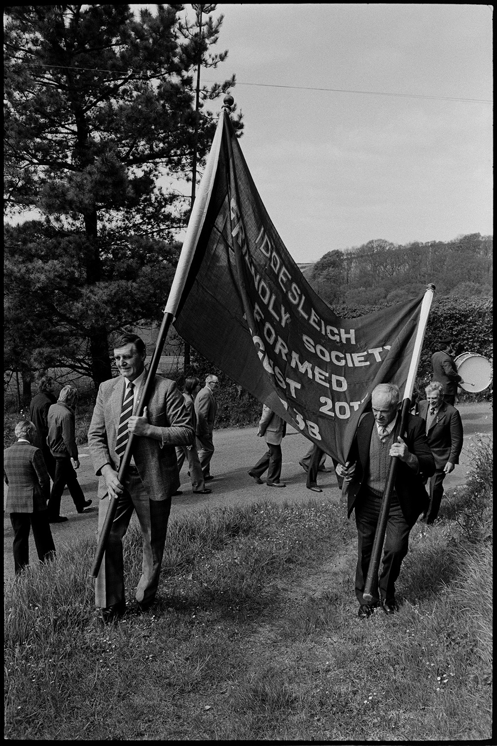 Club Day procession. Band and members marching to the Church. Entering church under banner.
[Men parading along a road and up the Iddesleigh church path in the Iddesleigh Club Day parade. The two men in the front are carrying the Iddesleigh Friendly Society banner.]