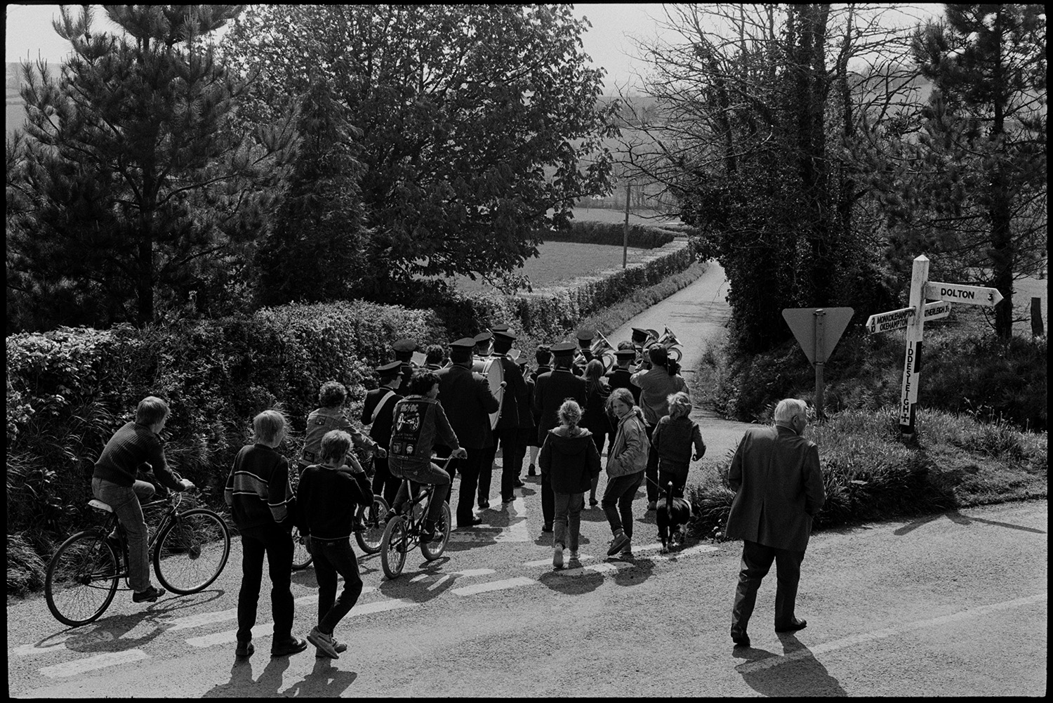 Club Day procession. Band and villagers marching to sports field.
[People, including children and boys riding bicycles, following the Hatherleigh Silver Band down a road in the Iddesleigh Club day parade. A signpost is at the road junction.]