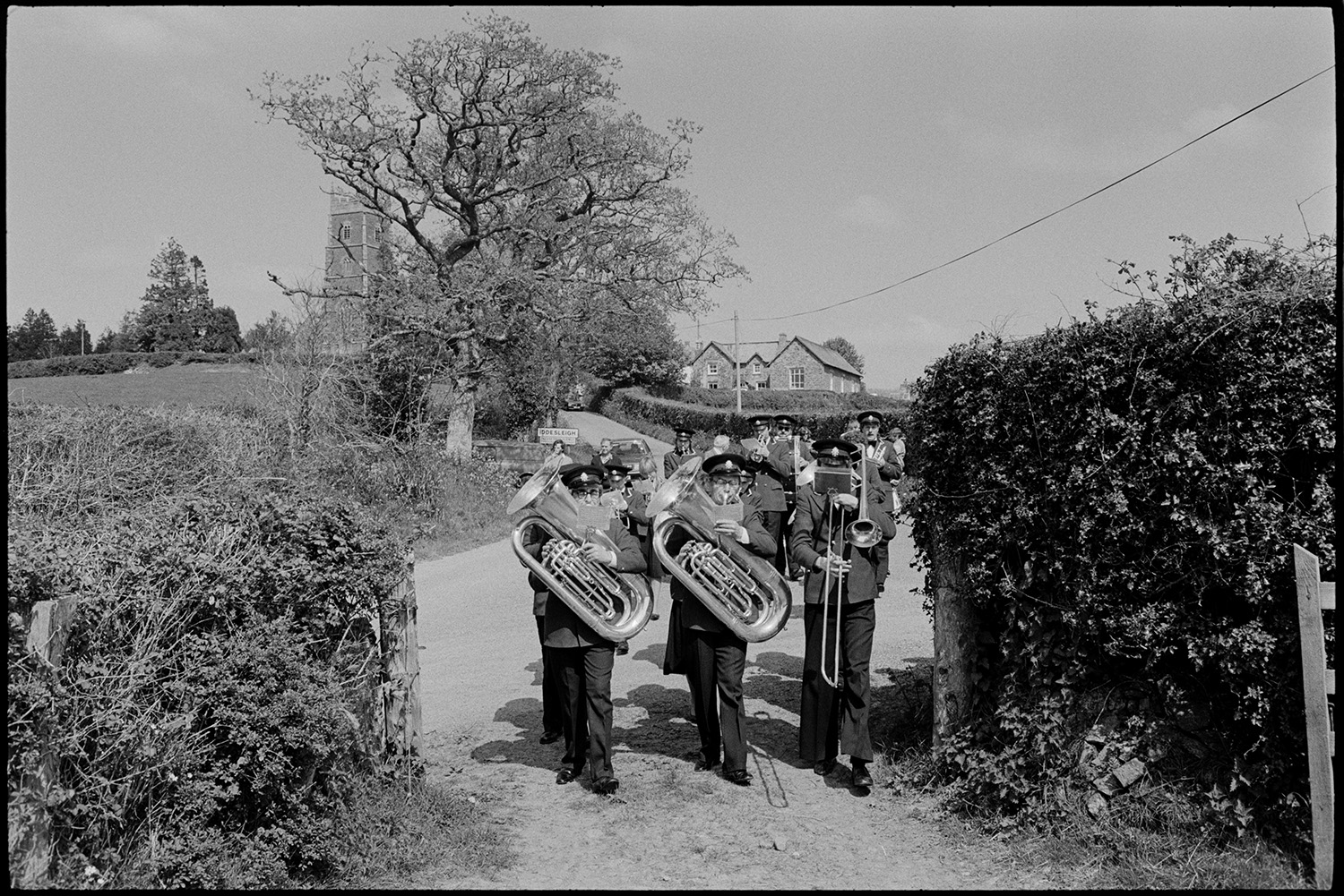 Club Day procession. Band and villagers marching to sports field.
[The Hatherleigh Silver Band parading down a road for the Iddesleigh Club Day procession.  Iddesleigh Church can be seen behind a tree in the background.]