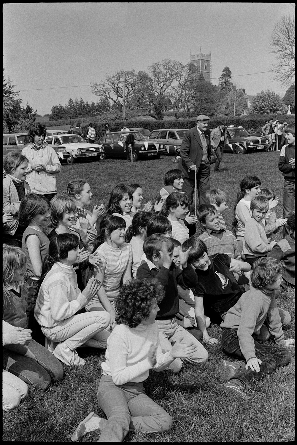 Club Day, sports, sack race, wellie throwing, man filming.
[A crowd of children and adults gathered in a field watching the sports at Iddesleigh Club Day. Parked cars and Iddesleigh Church tower can be seen in the background.]