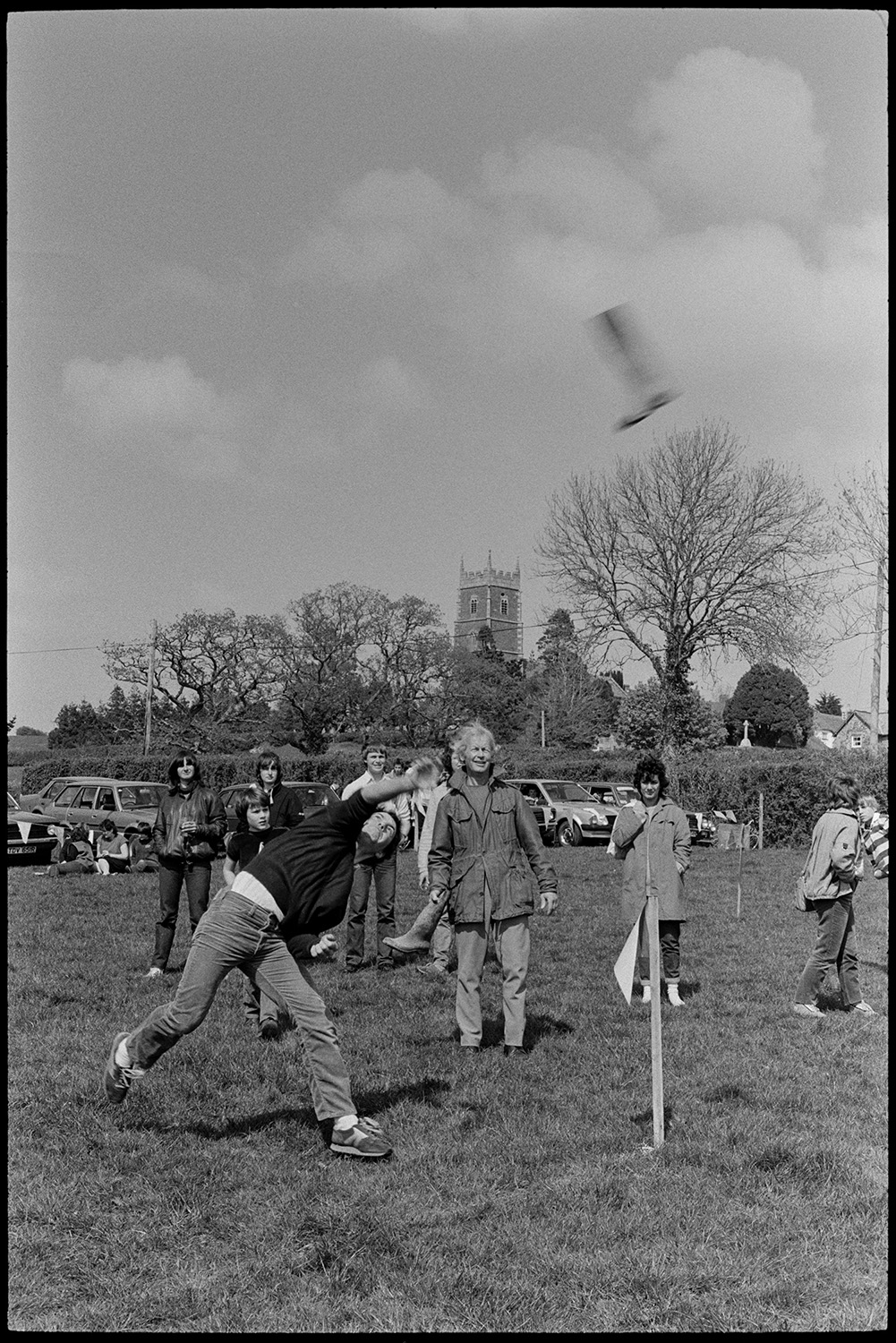 Club Day, sports, sack race, wellie throwing, man filming.
[People watching and taking part in wellie throwing in a field at Iddesleigh Club Day. Parked cars and Iddesleigh Church tower are visible in the background.]